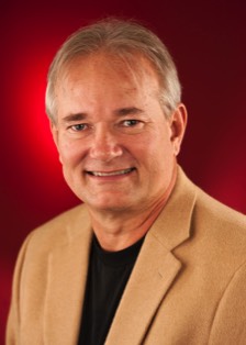 A color photo of Byron Bridges, a white man with grey hair, wearing a beige suit and black shirt smiling in-front of a red background.
