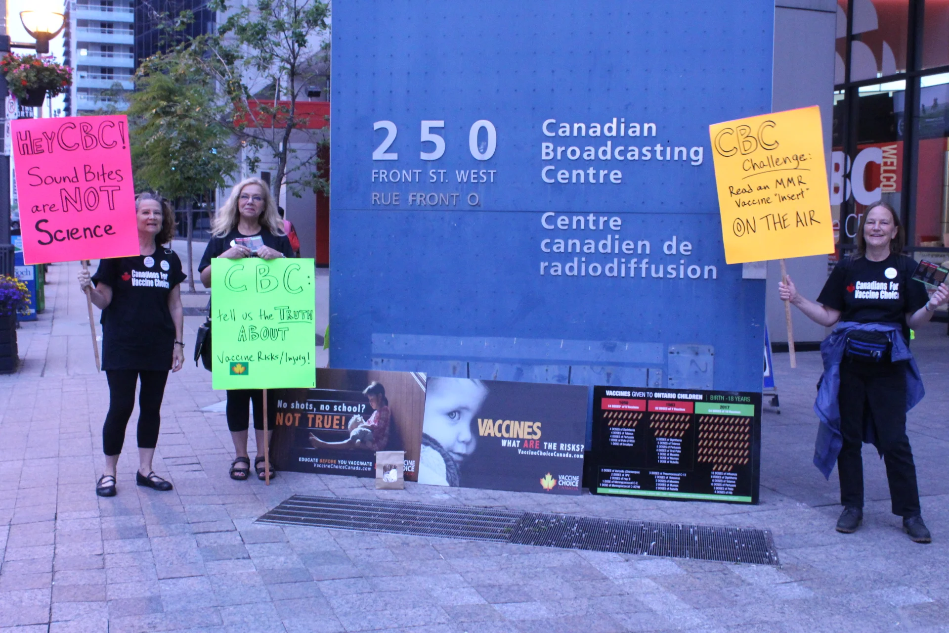Placards say, from left to right: Hey CBC. Sounds bites are not science; CBC: Tell us the truth about vaccine risks/injury; CBC Challenge: Read an MMR “insert” on the air.