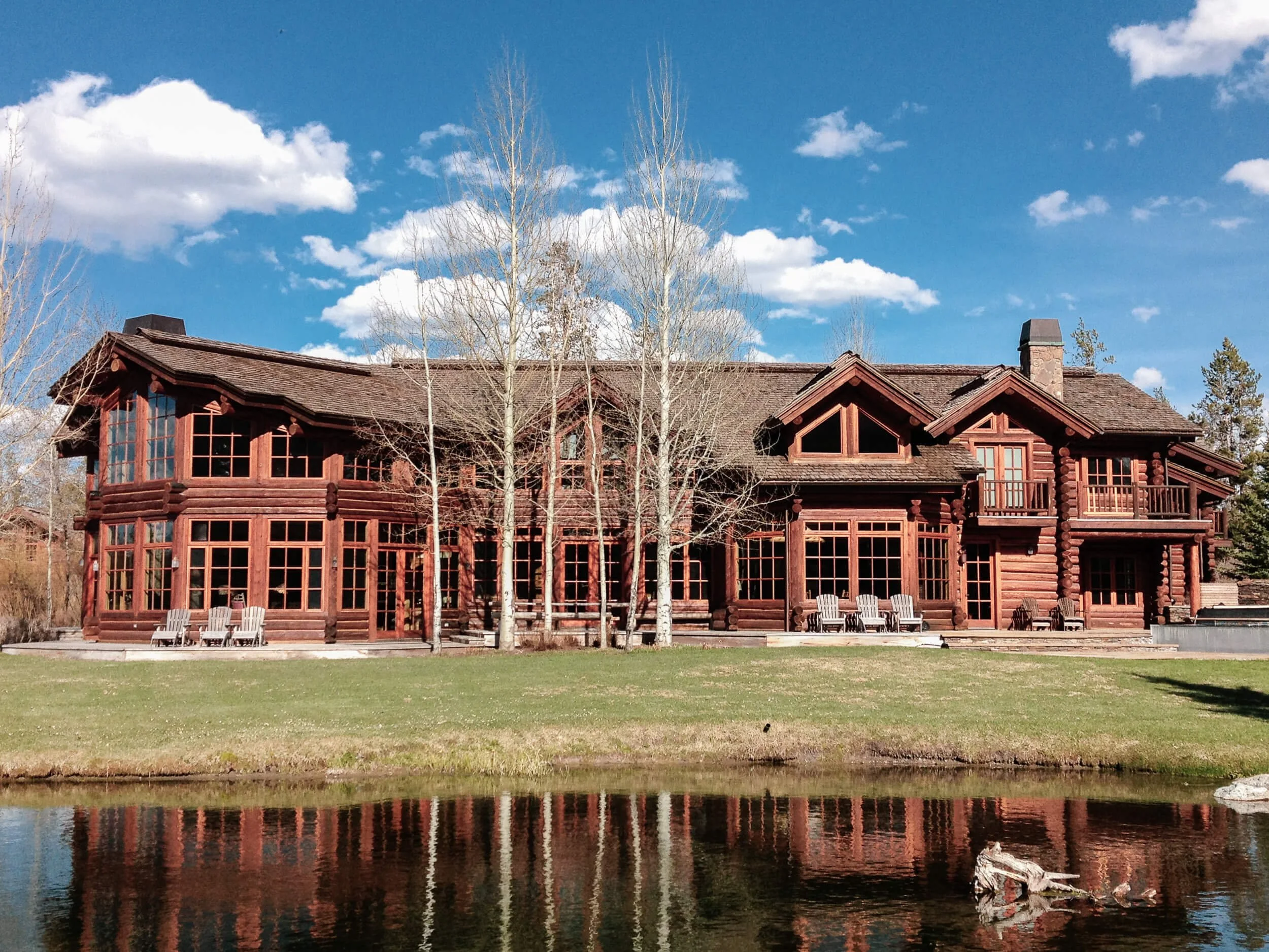 Large timber-frame home with expansive windows and a balcony, reflected in the pond below under a partly cloudy sky.