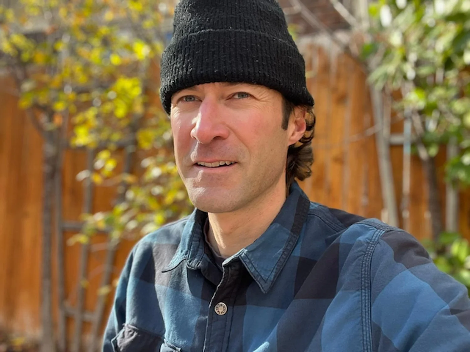 A man wearing a black beanie and a blue plaid shirt taking a selfie outdoors with a background of a wooden fence and green foliage.