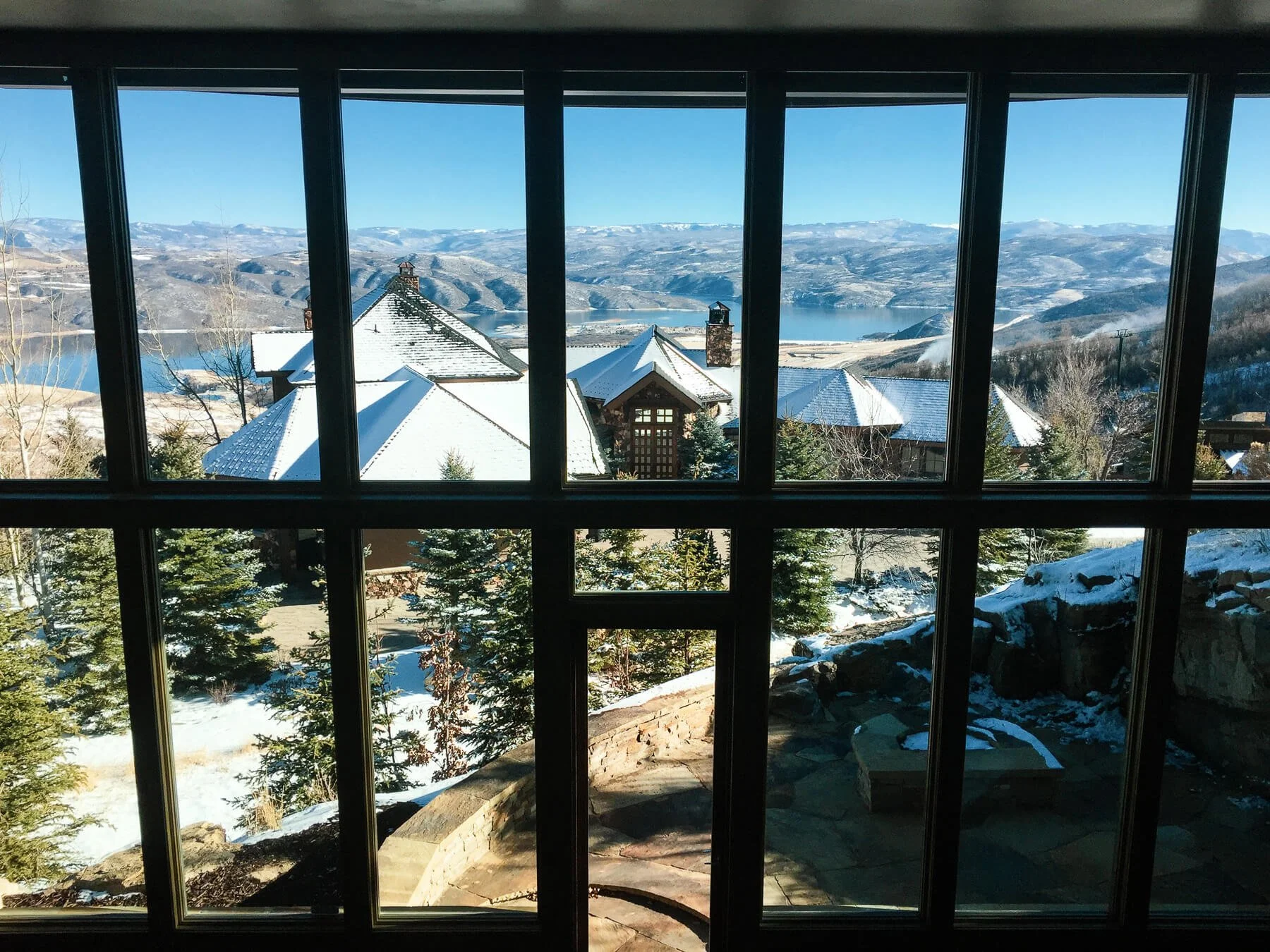 Snow-covered rooftops and a landscape of mountains and a lake viewed through a window with multiple panes. Pine trees and rocks are in the foreground.