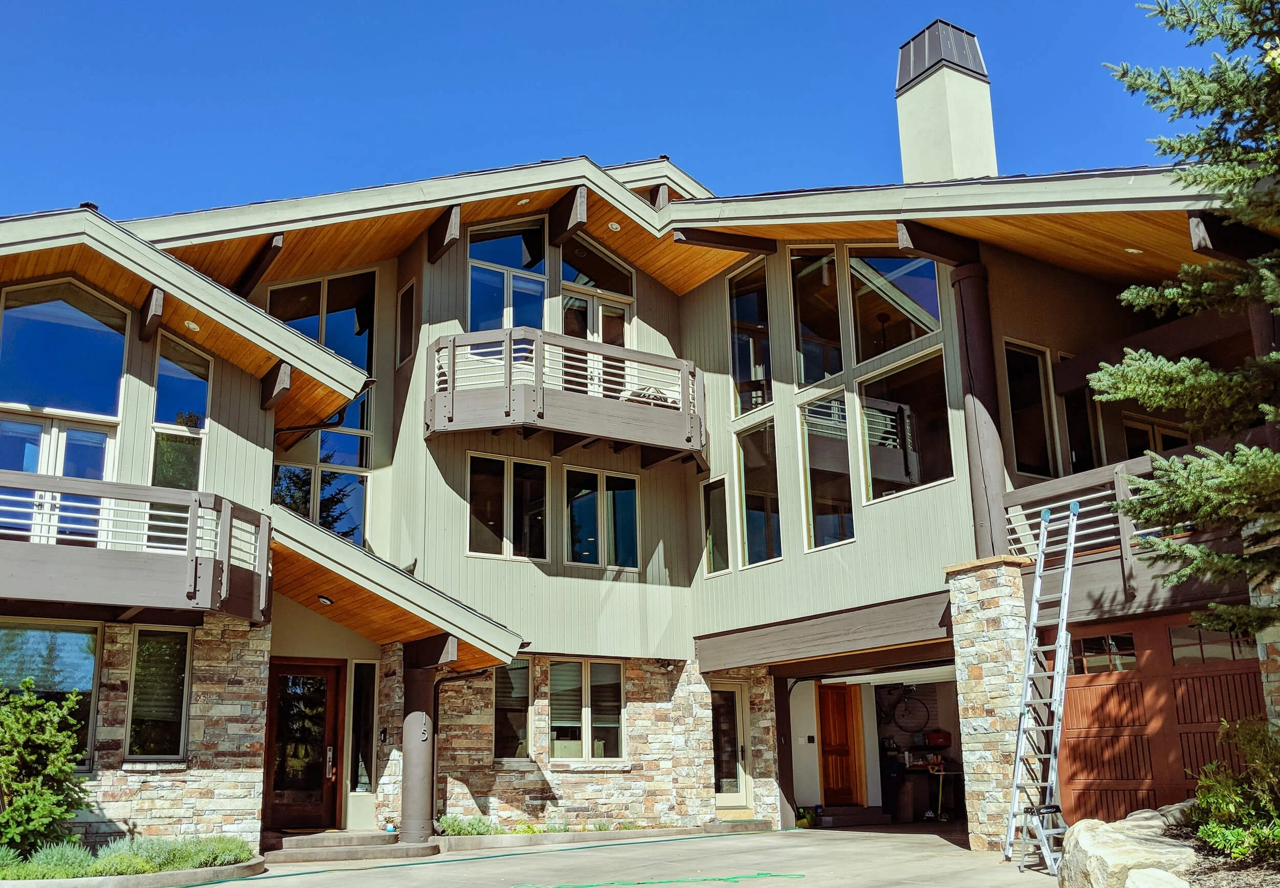 A modern multi-story house with large, angular windows, a balcony, stone accents, and a chimney under a clear blue sky. A ladder leans against the house on the right side, and some greenery is visible in the foreground.