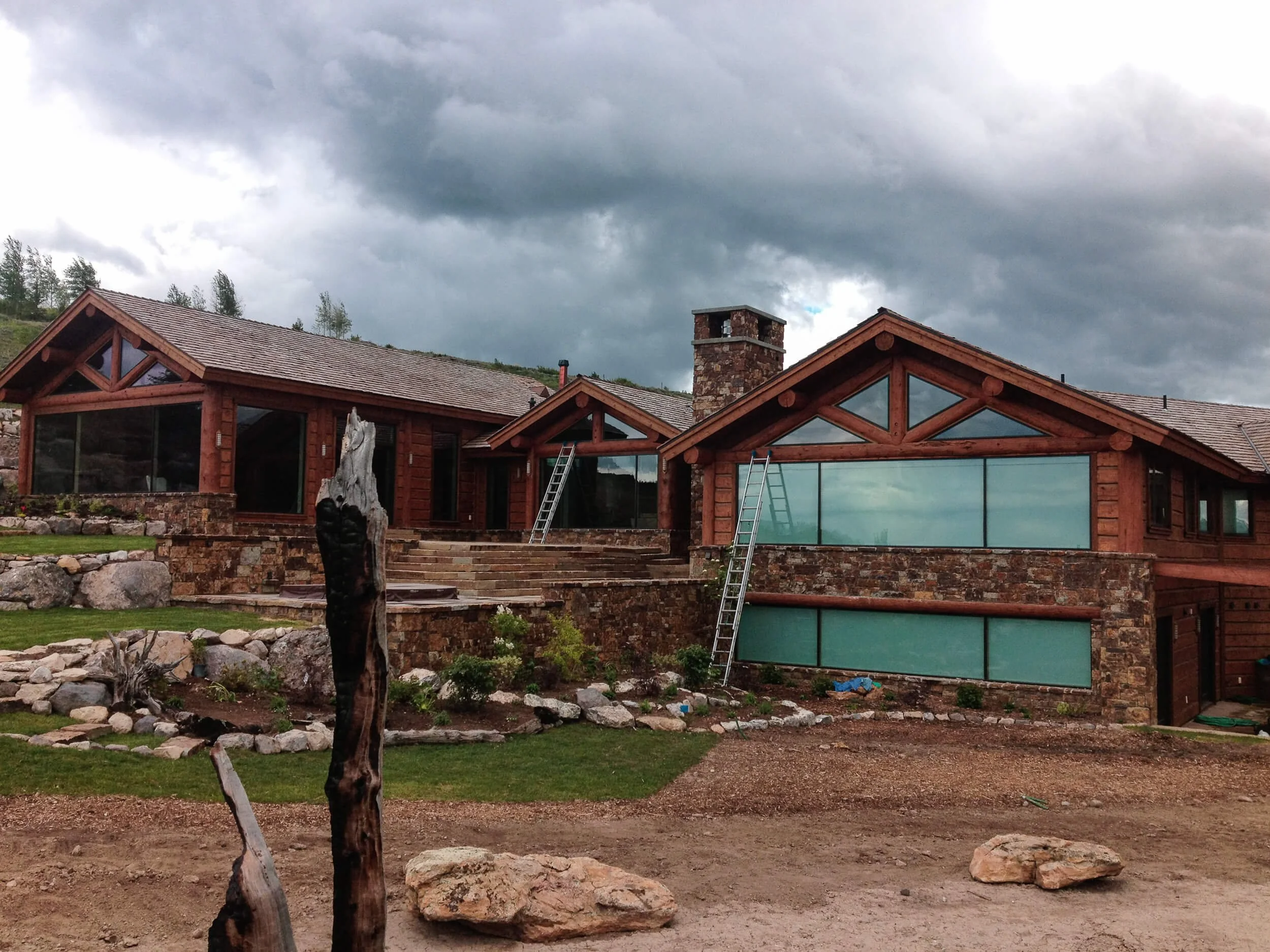 Mountain home under construction, showing its wood frame, stone chimney, and large glass windows being installed.