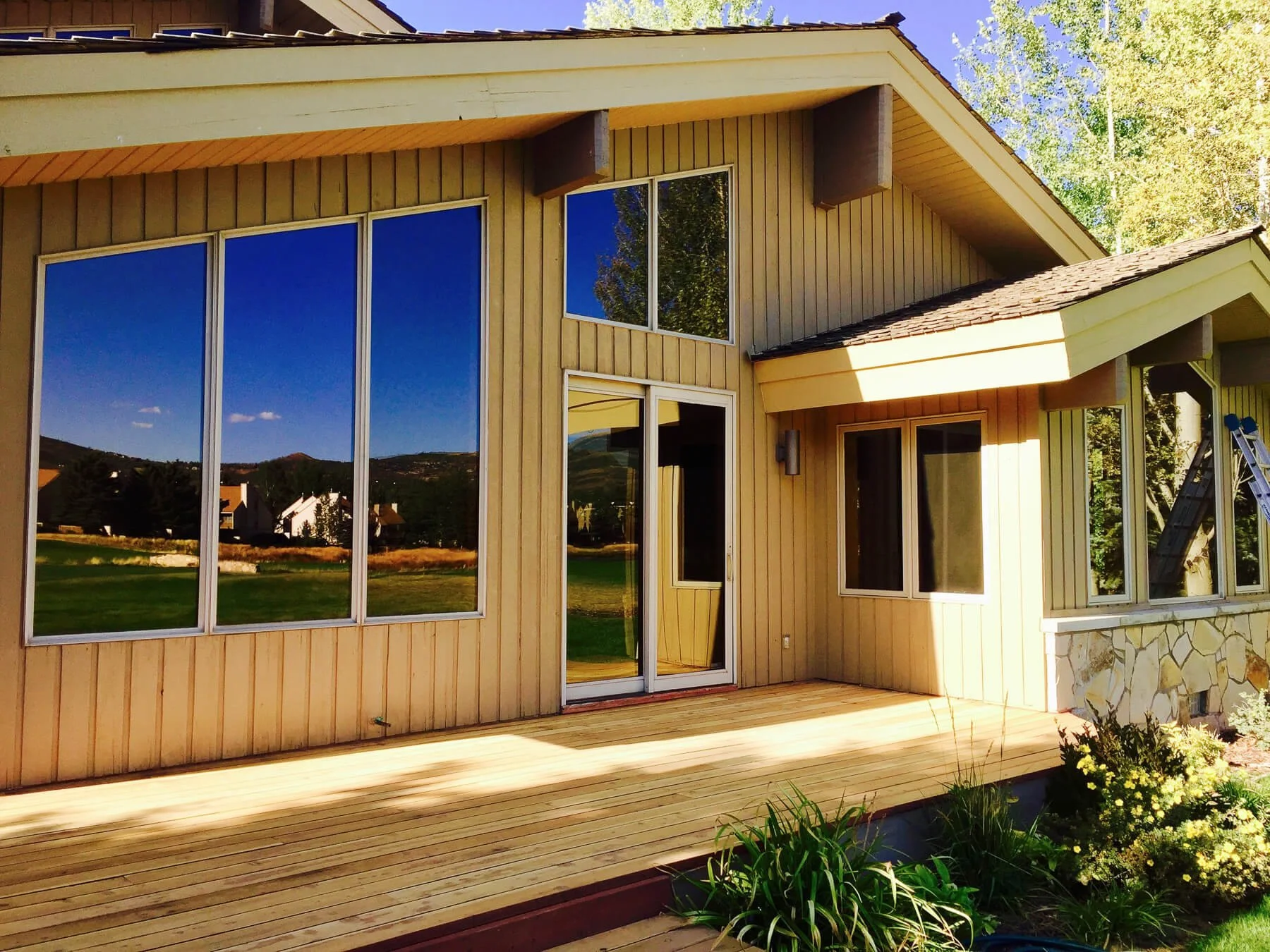 Freshly painted yellow house with large glass windows reflecting the blue sky and green landscape, wooden deck, and garden with green plants and yellow flowers in the foreground.