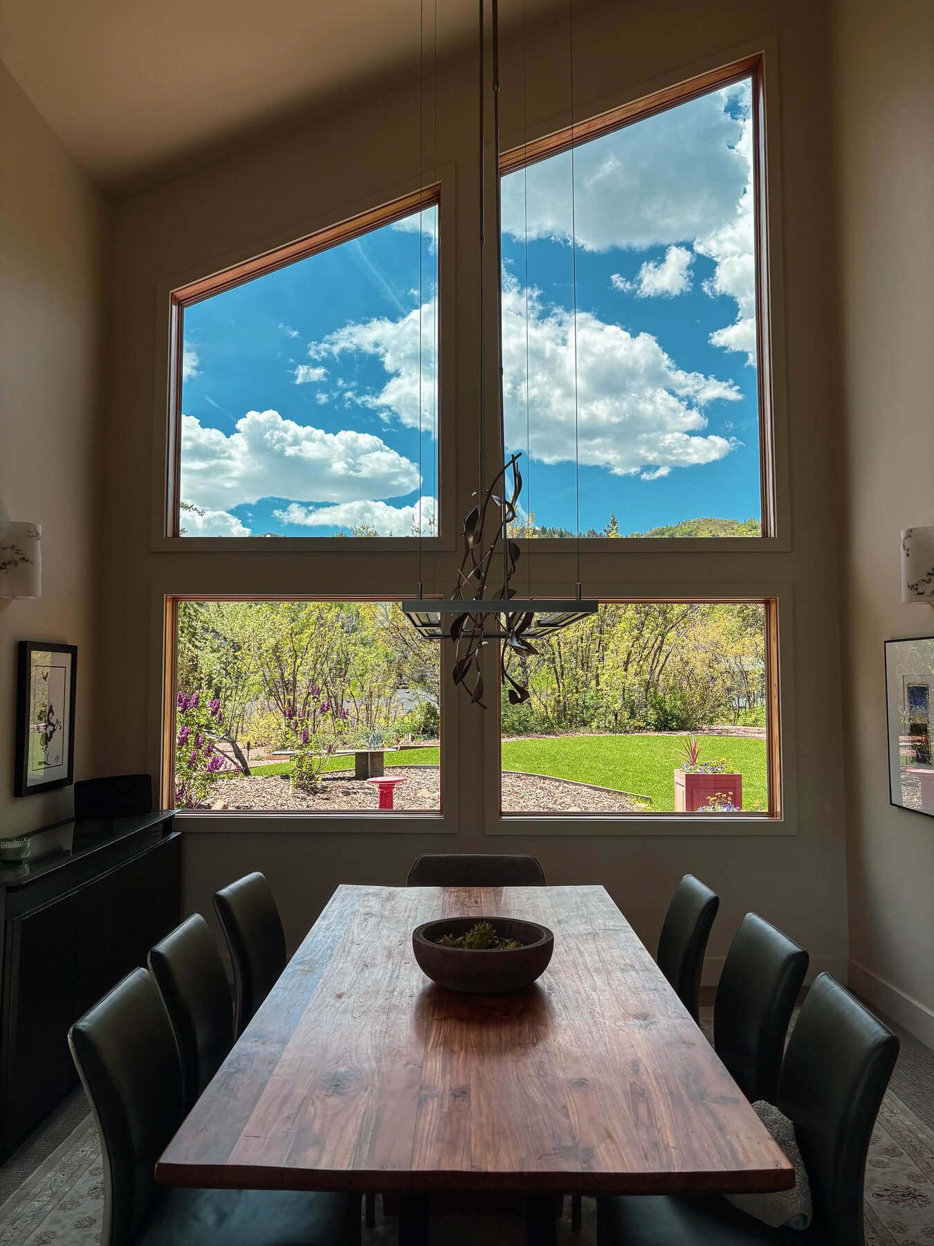 A dining room with a large wooden table surrounded by black chairs, and a bowl on the table. Large windows reveal a garden with trees, purple flowers, and a lawn under a blue sky with clouds.