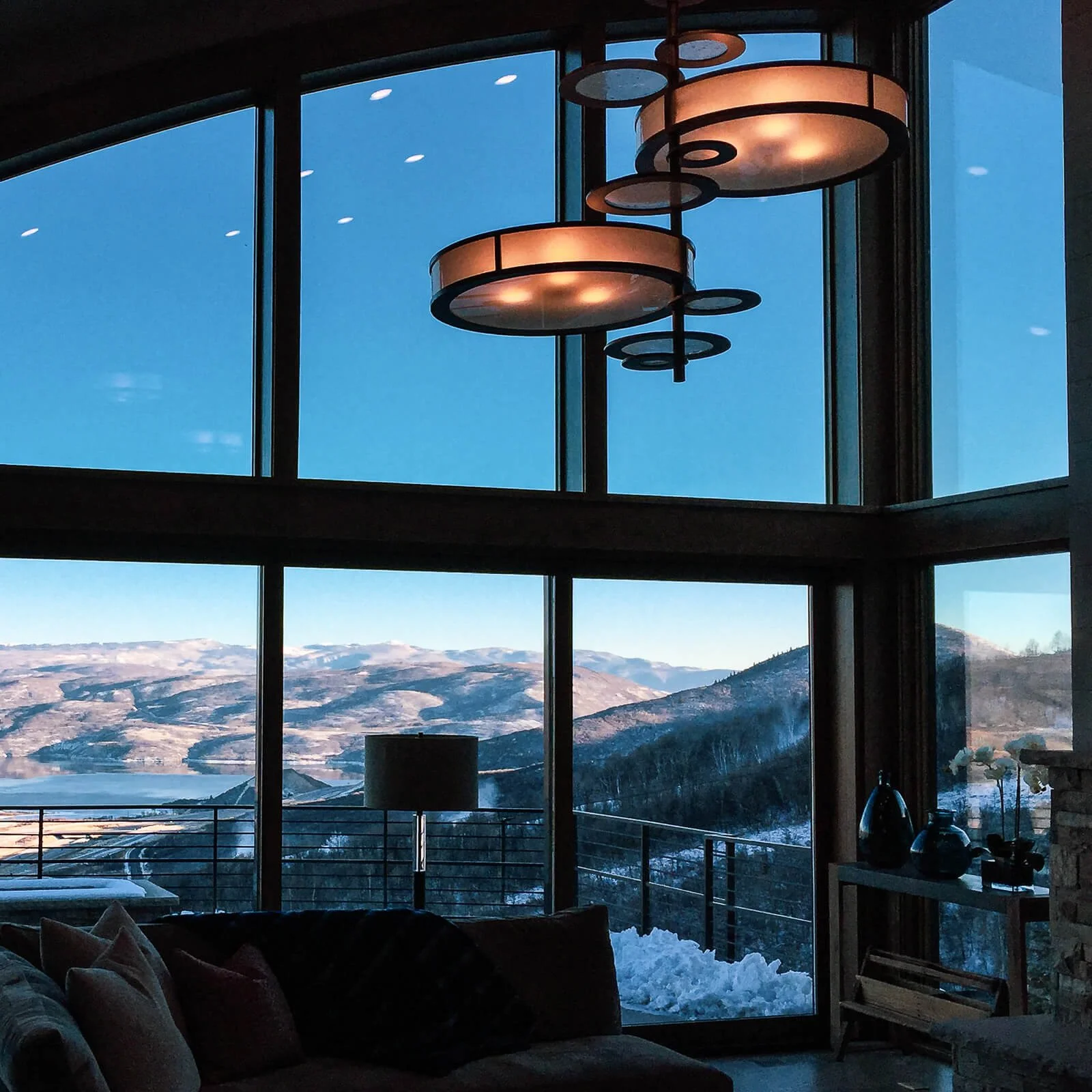 Living room with floor-to-ceiling windows overlooking snow-capped mountains, pendant lighting, and a sofa with pillows.