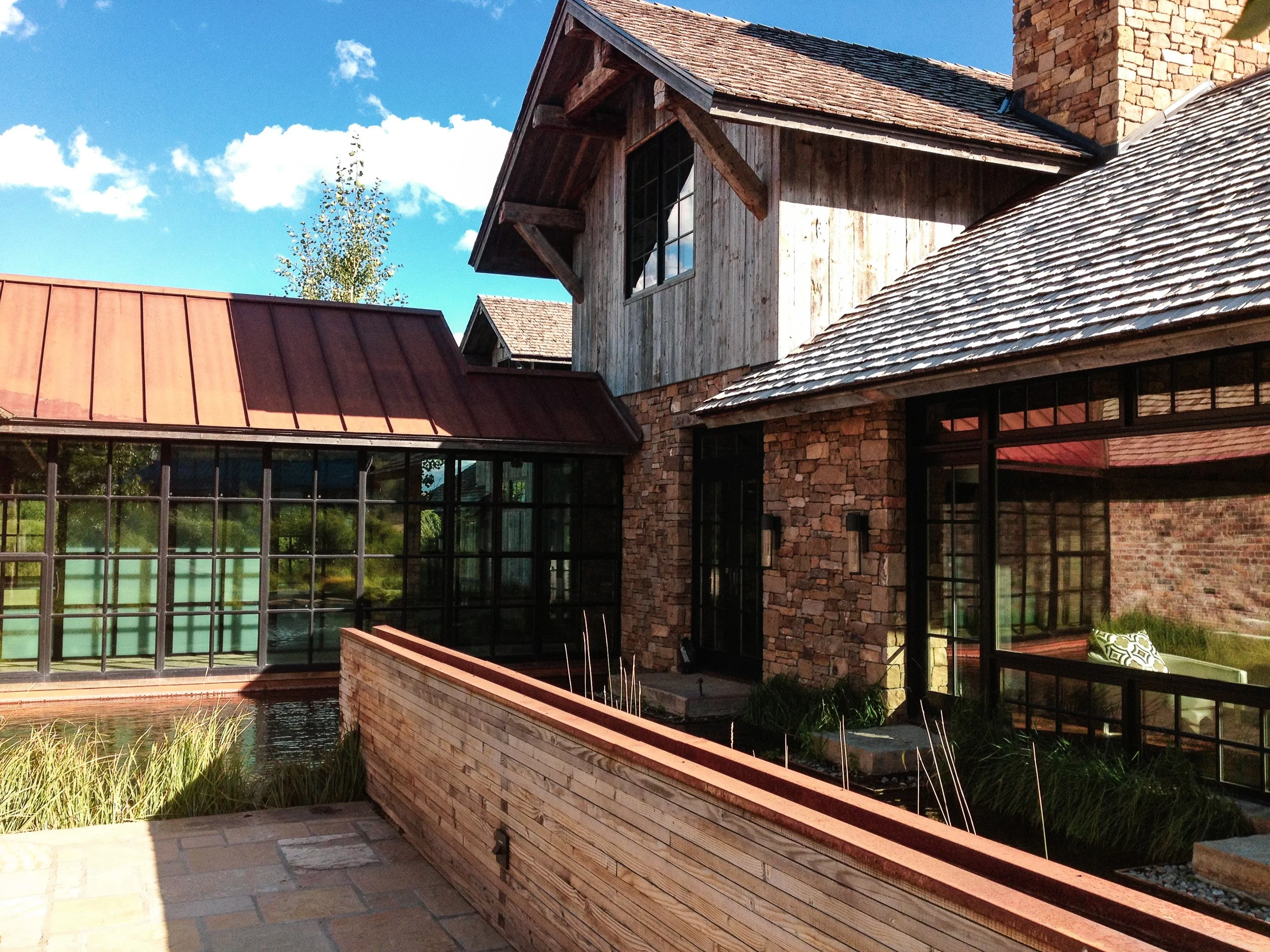 A rustic house with a sloped roof covered in shingles, a stone chimney, and large windows, set against a bright blue sky with white clouds.