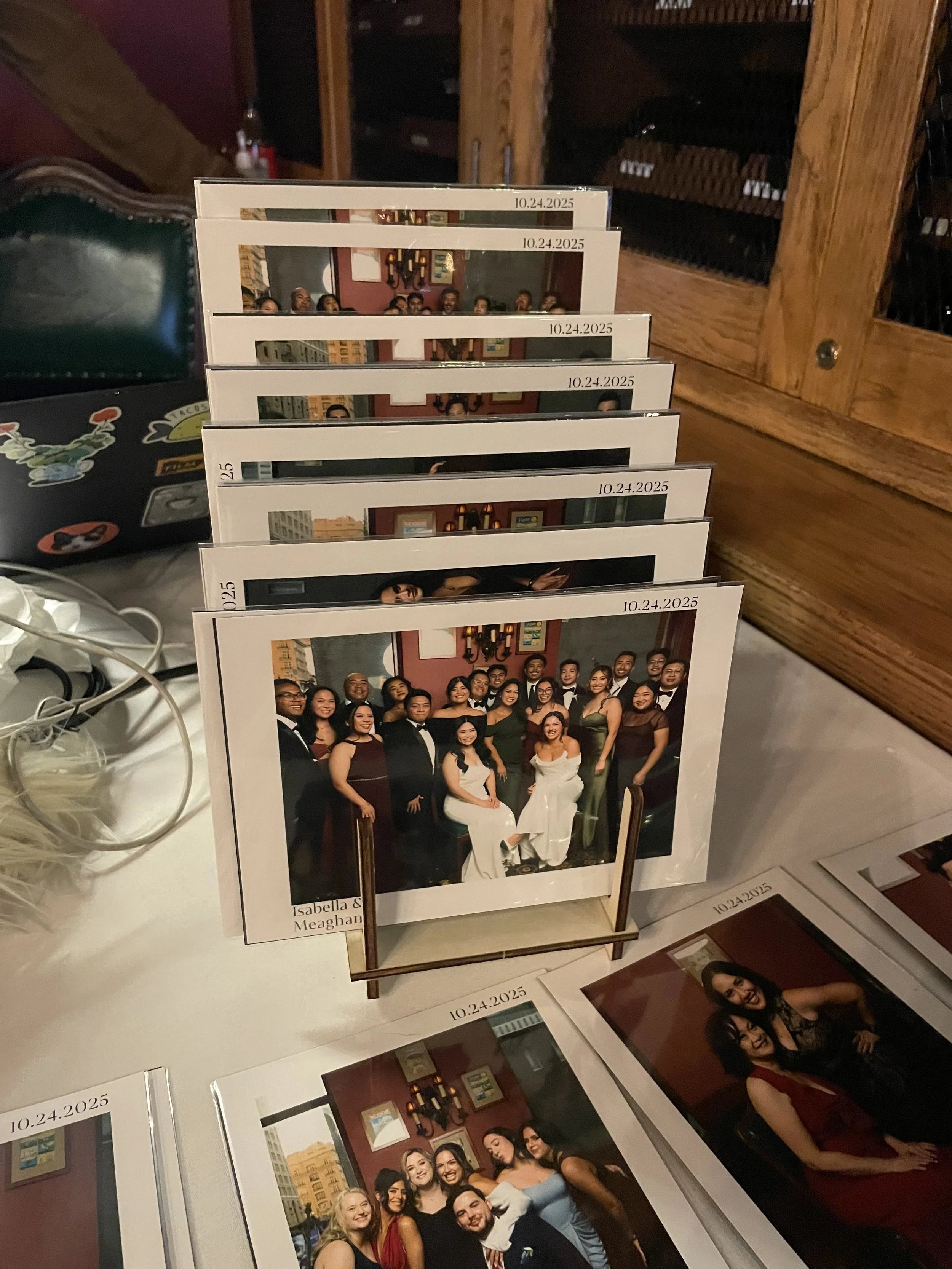 A collection of printed photographs of people at a wedding in one of those portrait station lounges, displayed on a stand. The top photo shows a large group of people dressed in formal attire, with a date printed on it for the date of event