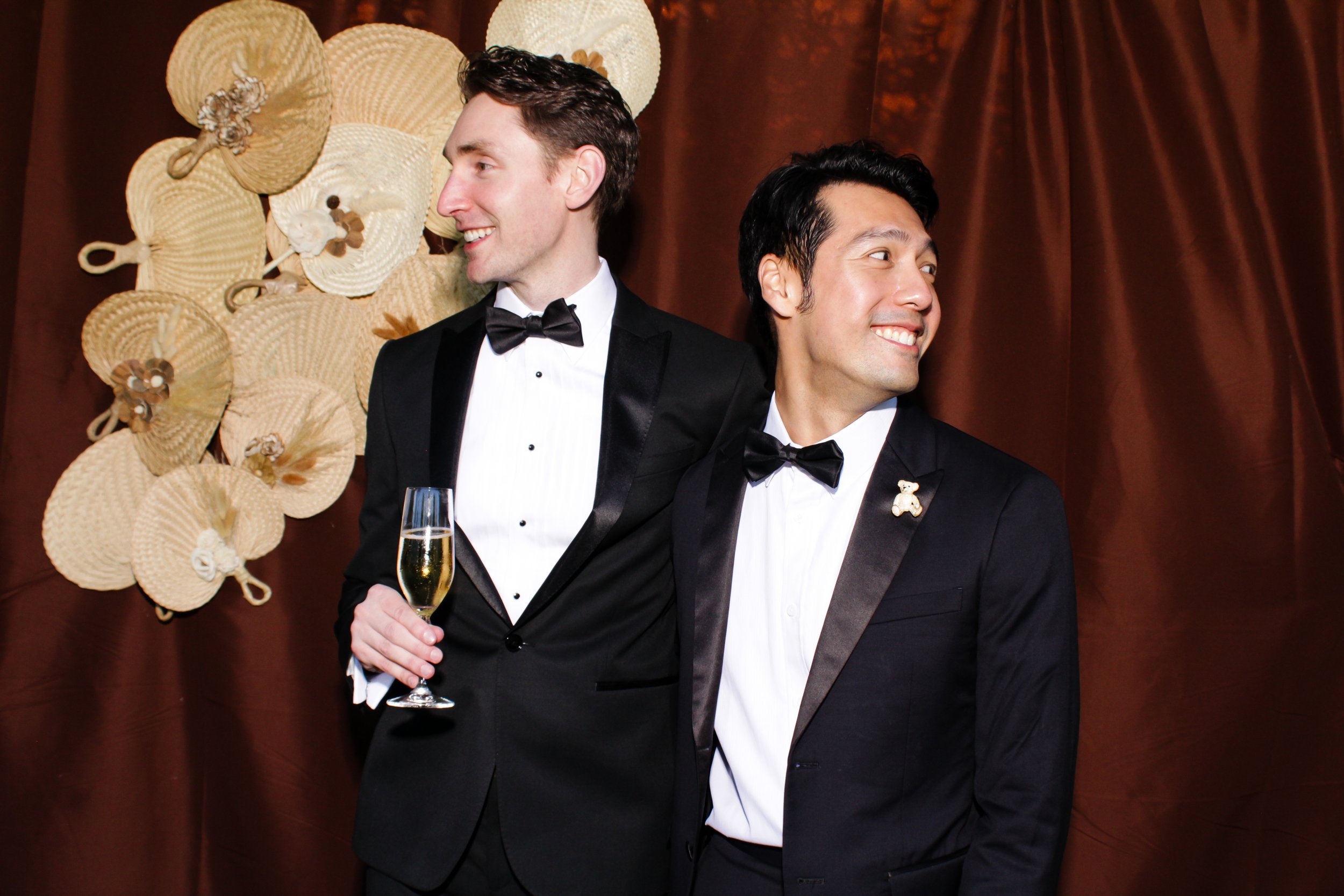 Two men dressed in tuxedos in an outdoor open-air photo booth, one holding a glass of champagne, standing in front of a custom designed backdrop decorated with woven fans from the Philippines and burlap flowers.