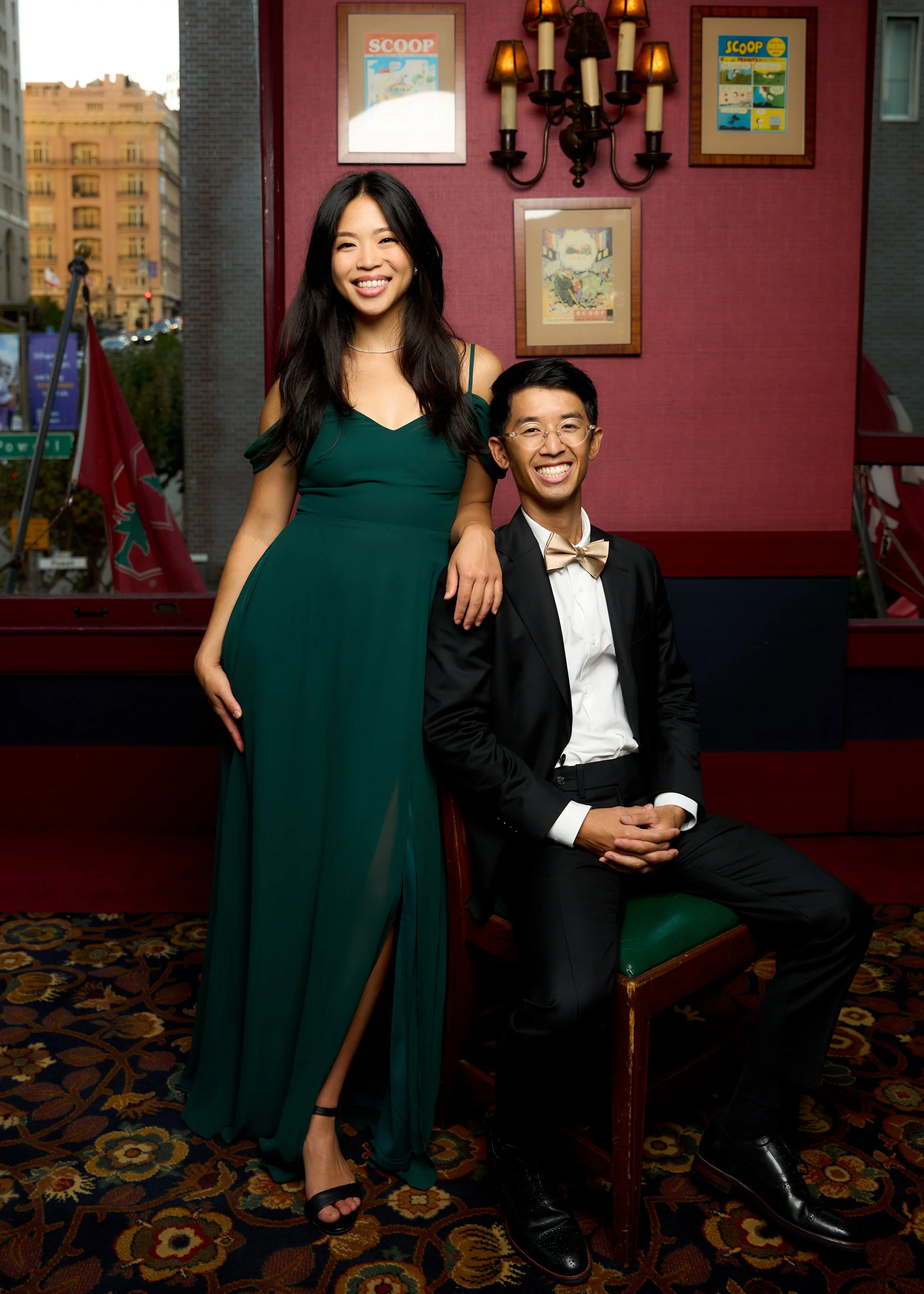 A young woman in a green dress and black high heels standing next to a seated young man in a tuxedo with a bow tie, smiling together in a decorated indoor setting with framed artwork and wall sconce lights.