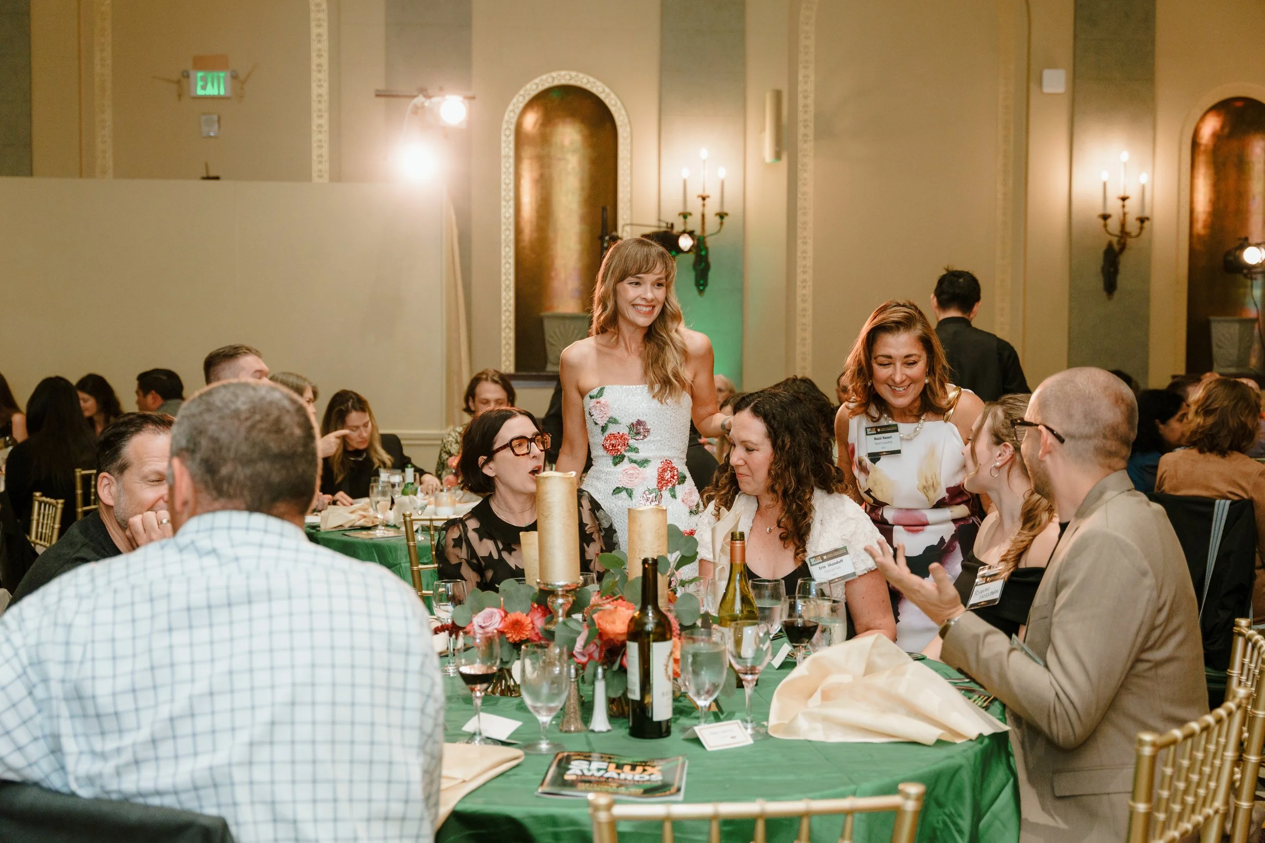 A group of people at a formal dinner event, with a woman in a floral dress standing and smiling among seated guests in a decorated banquet hall.