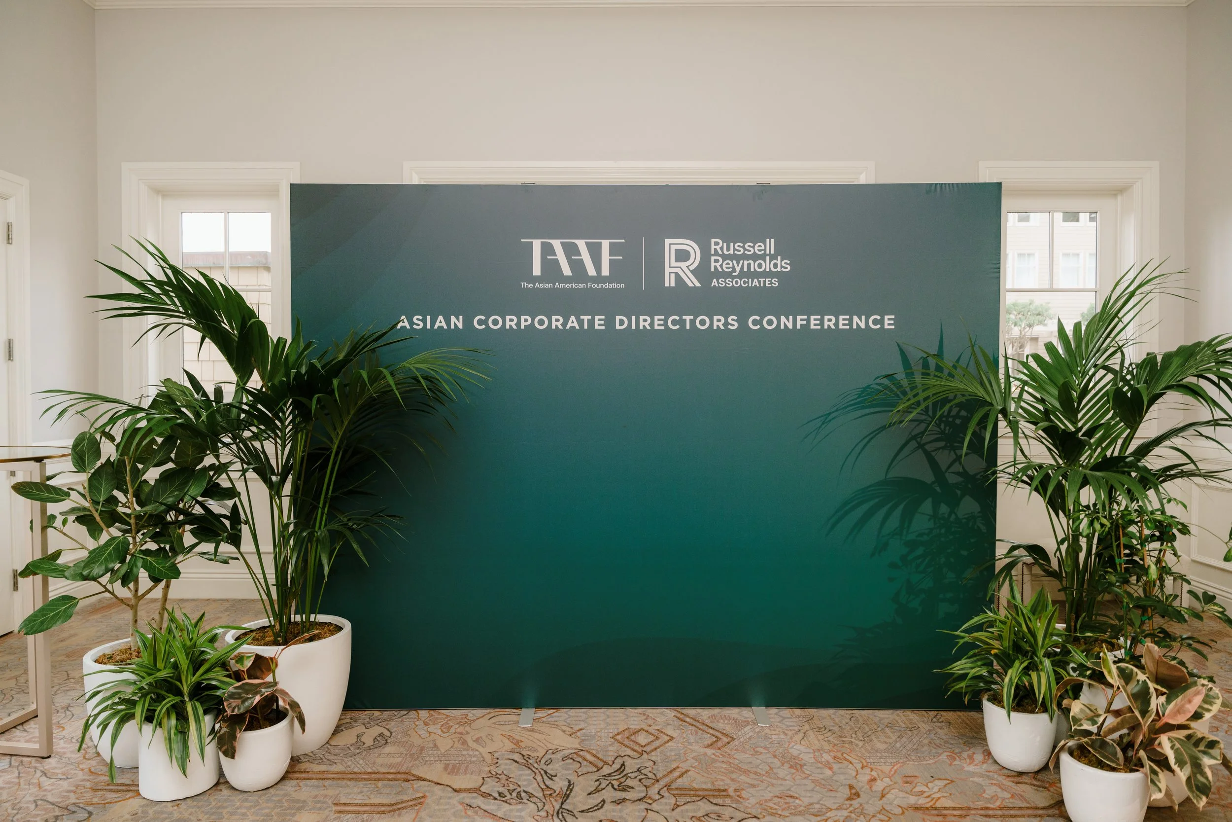 A large green backdrop with the logos of the Asian American Foundation and Russell Reynolds Associates, titled "Asian Corporate Directors Conference," is surrounded by potted green plants on a patterned carpet near windows in a well-lit room.