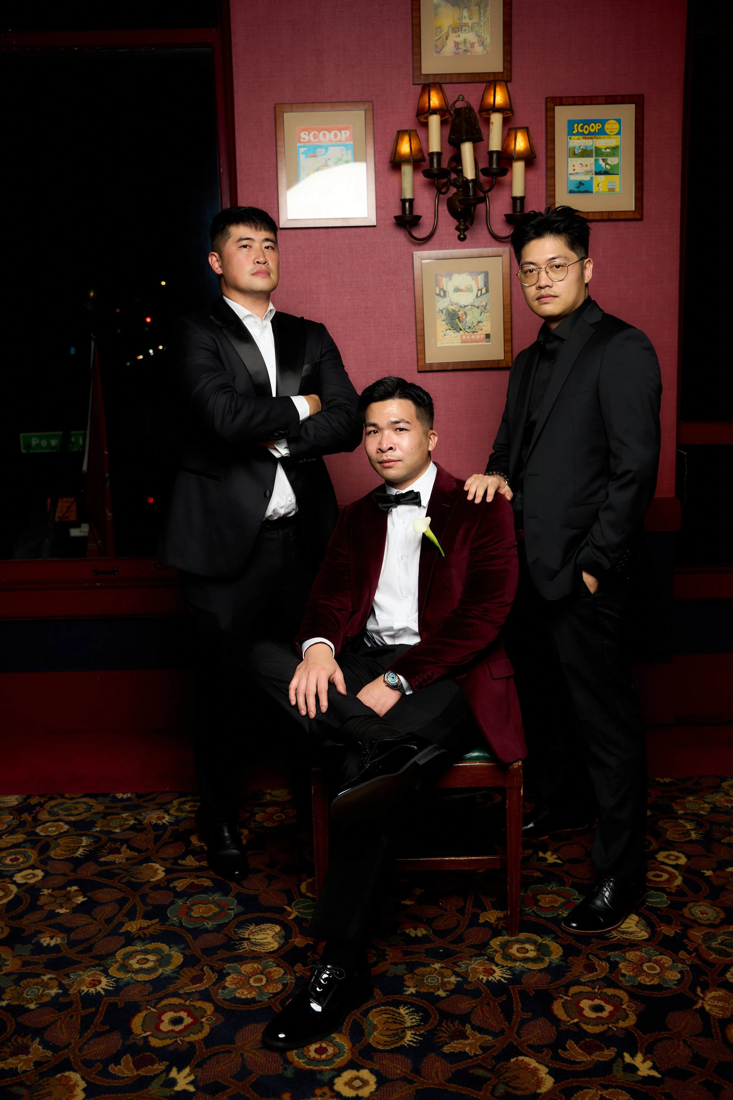 Three young men dressed in formal wear posing in a Six & Ten Studios onsite portrait studio at a wedding in San Francisco located in a room with framed artwork and a chandelier on a red wall, with patterned carpet on the floor.