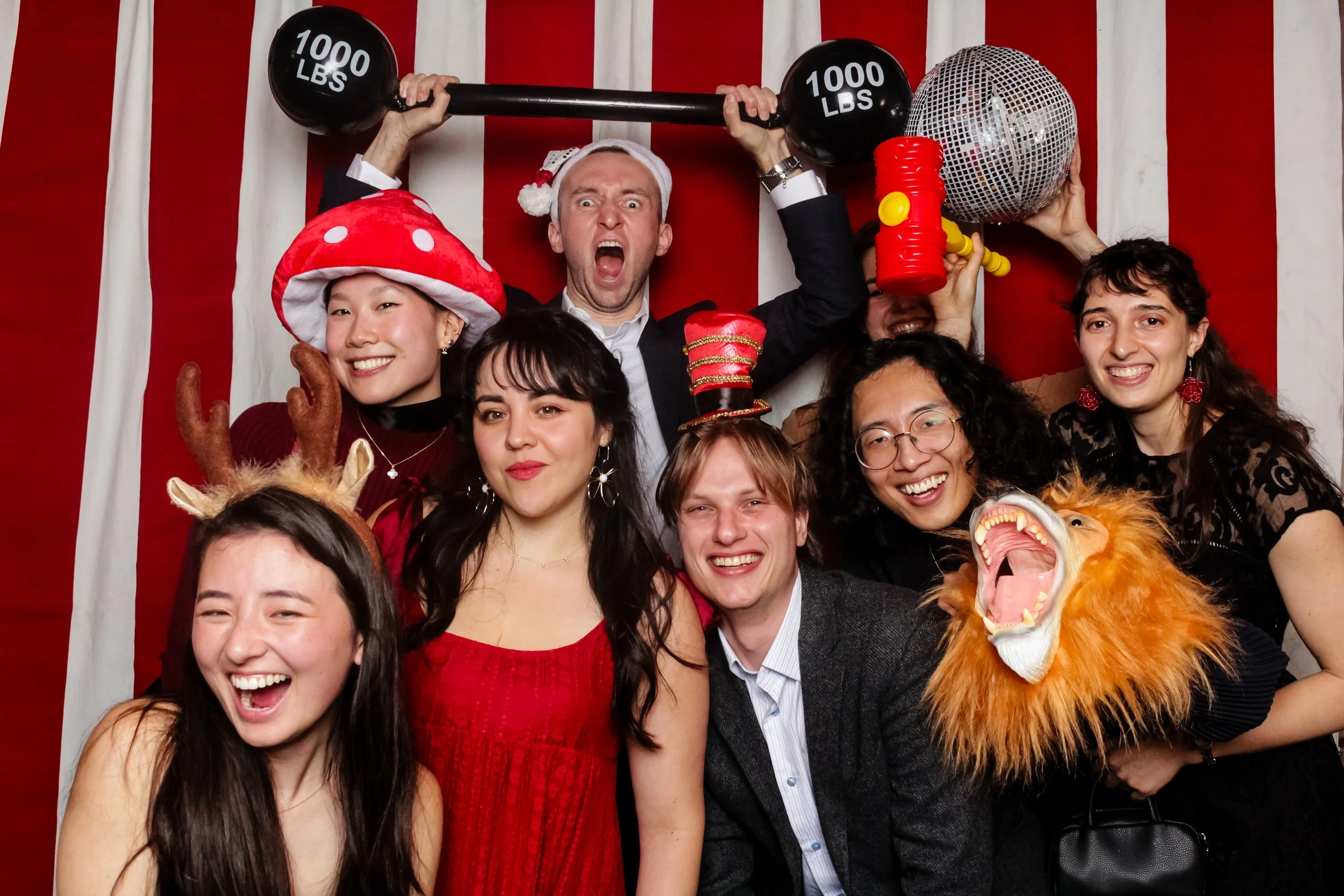 A group of people celebrating in front of a red and white striped background, wearing festive costumes and accessories. Some are holding props like a barbell marked 1000 lbs, a disco ball, and a lion mask in an open-air photo booth in San Francisco.