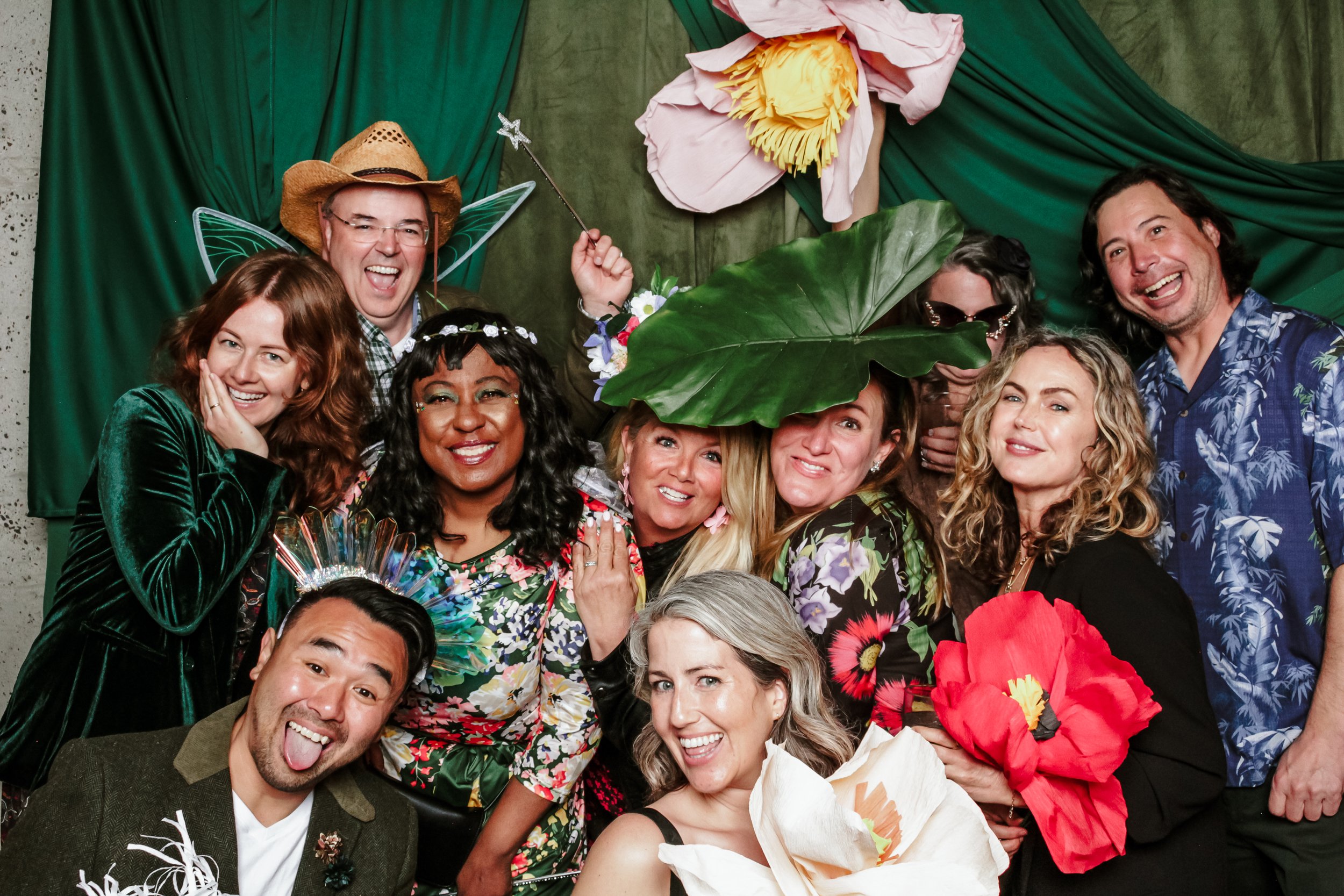 Group of people taking photos in a photo booth celebrating with custom props, including large flowers, leaves, and fairy wings, against a custom floral & green backdrop.