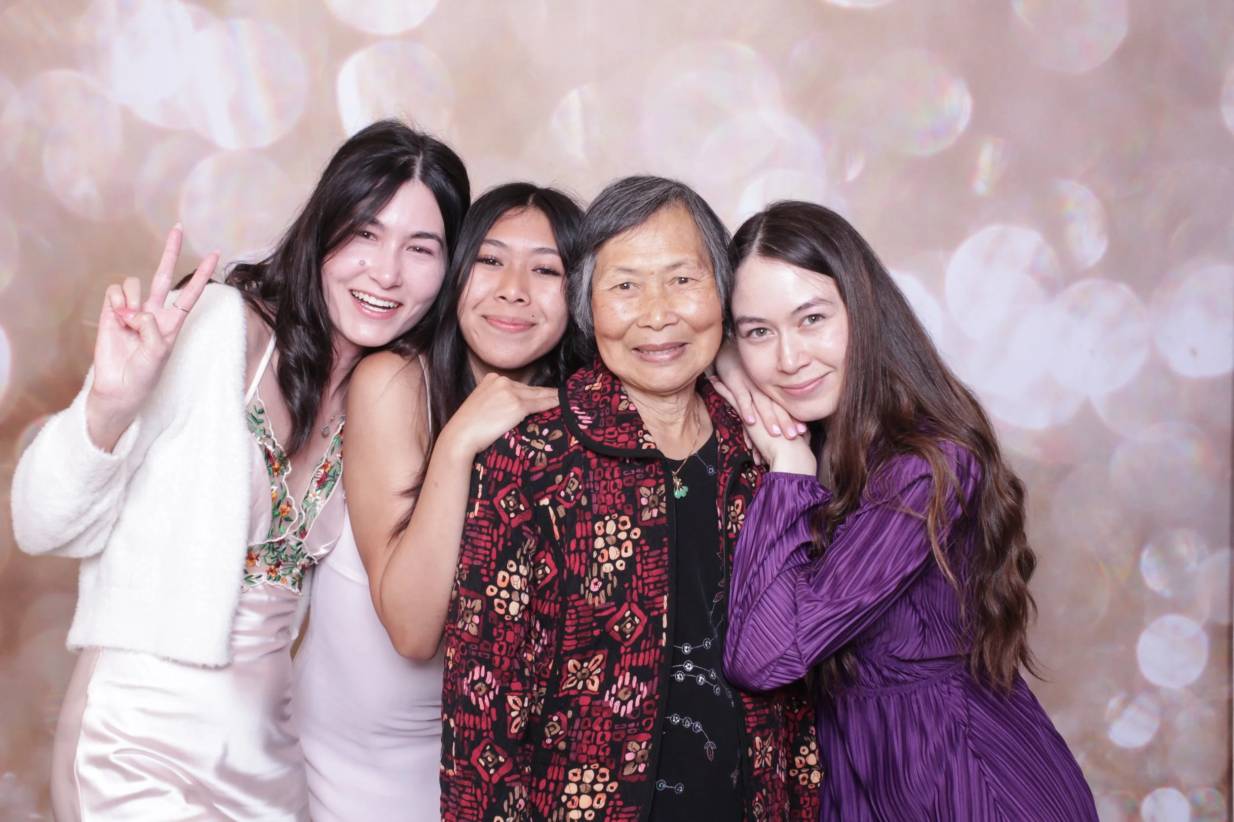 Group of four women and one older woman smiling and posing together in an open-air photo booth against a sparkly, bokeh background, with one woman showing a peace sign.