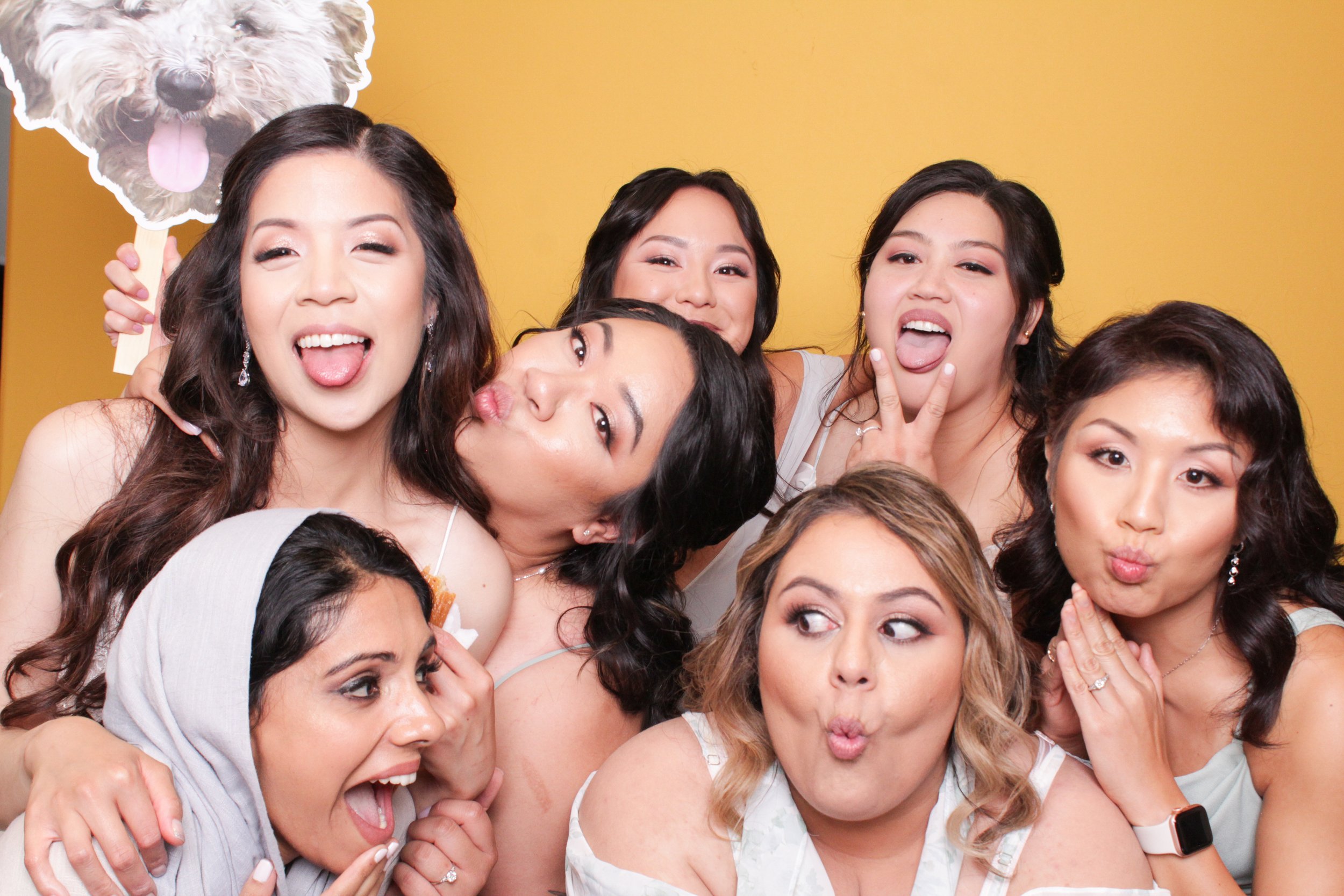 Group of women making playful faces and gestures in an open-air modern photo booth in front of a yellow background, with one holding a cutout custom prop of a dog.