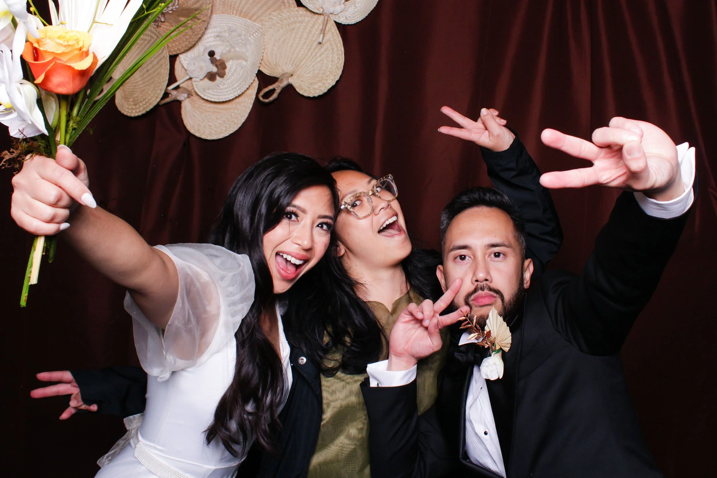Three people celebrating, smiling, making peace signs, with a woman holding a bouquet, a dark red curtain backdrop, and straw hats hanging on the wall.