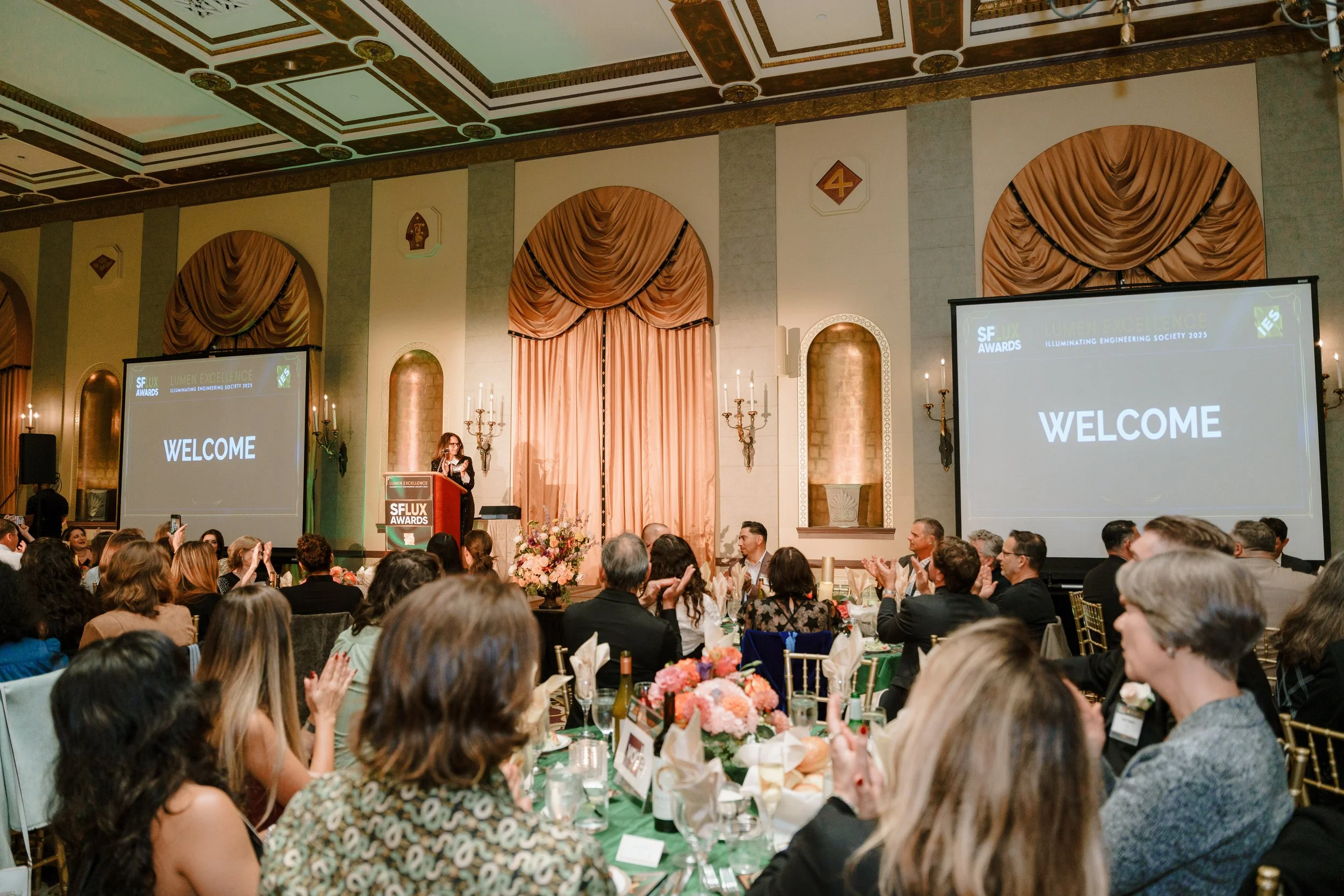 An indoor awards ceremony with a large audience seated at decorated tables, a stage with two screens displaying 'Welcome' and the event title 'San Francisco Lux Awards 2025', and a woman speaking at a podium.