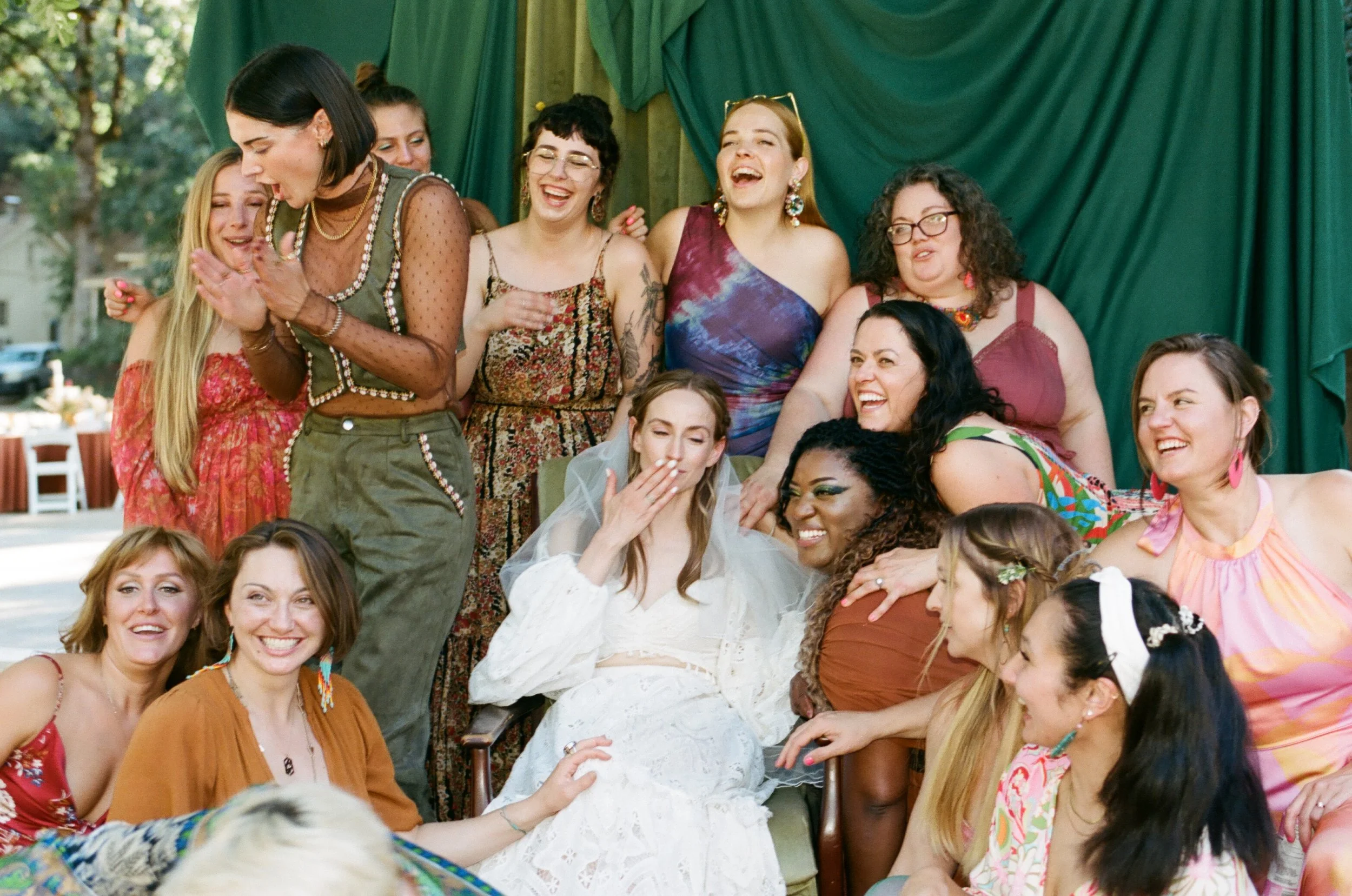 Group of women celebrating together at a wedding event, hugging and smiling, with green drapery curtains in the background in an outdoor mobile photography studio portrait lounge