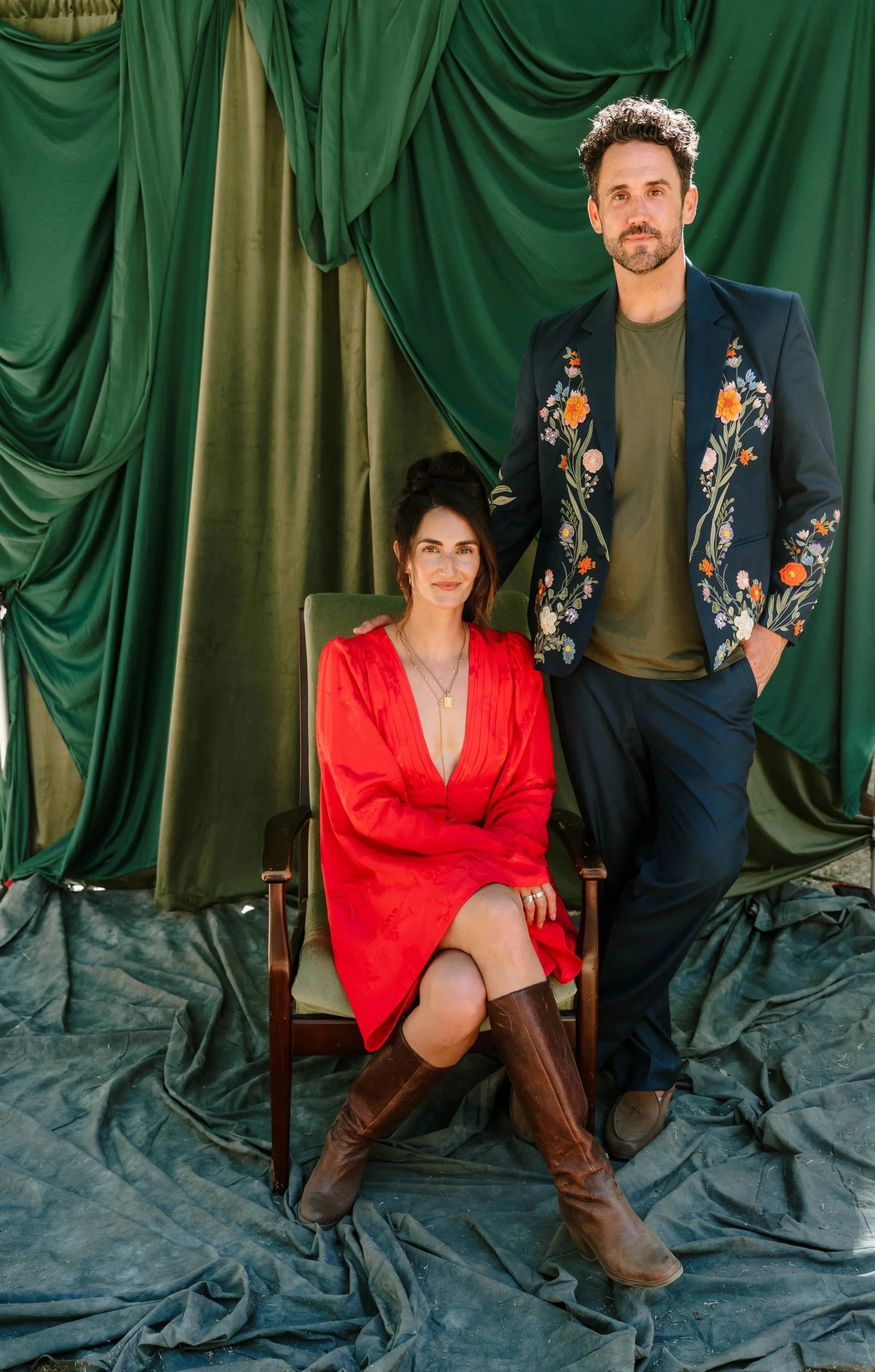 A woman in a red dress and brown boots sits on a green chair, and a man in a floral embroidered blazer and green shirt stands behind her with his hand on her shoulder, against a backdrop of green draped fabric in an outdoor portrait lounge experience
