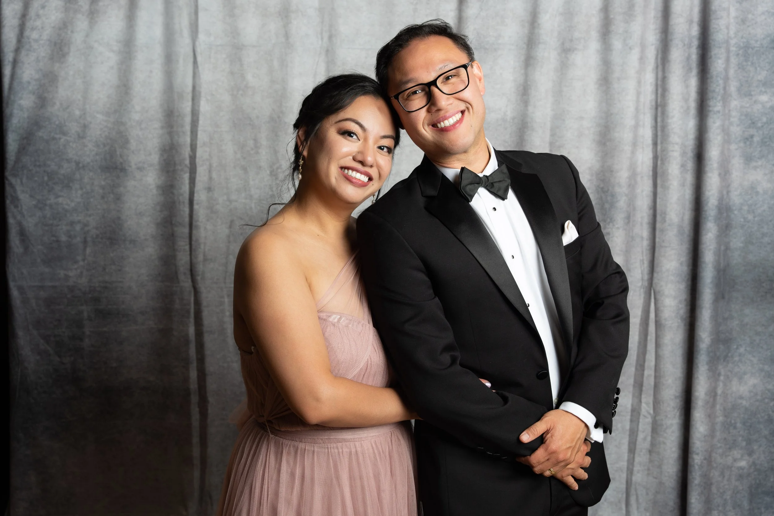 A smiling couple dressed in formal attire, standing close together in front of a gray velvet background in Six & Ten Studios onsite portrait booth in San Francisco Bay area.
