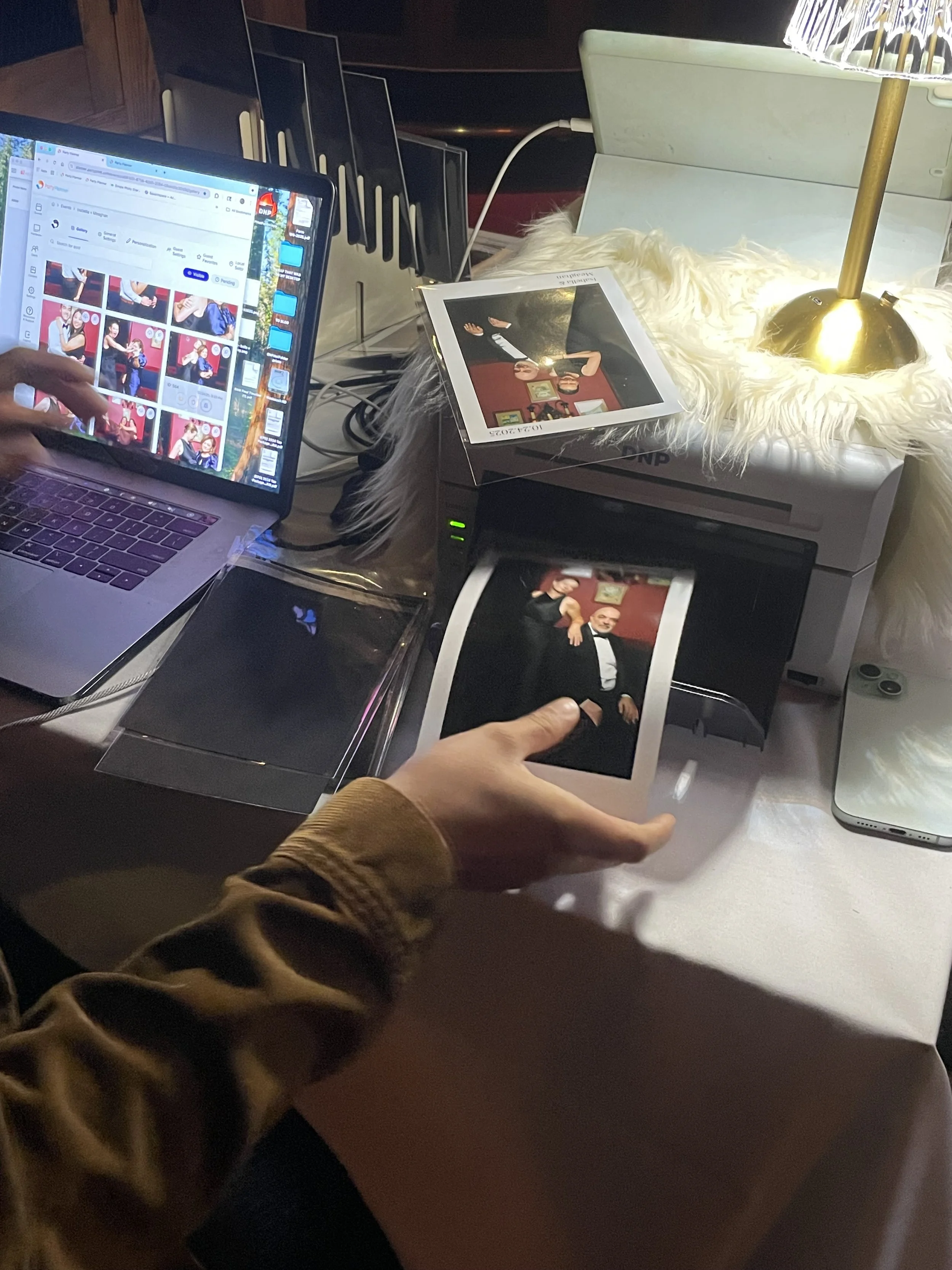 A workspace of the portrait lounge with laptops, printed photographs, a desk lamp with a gold base and white shade, and various electronic accessories on a white desk.