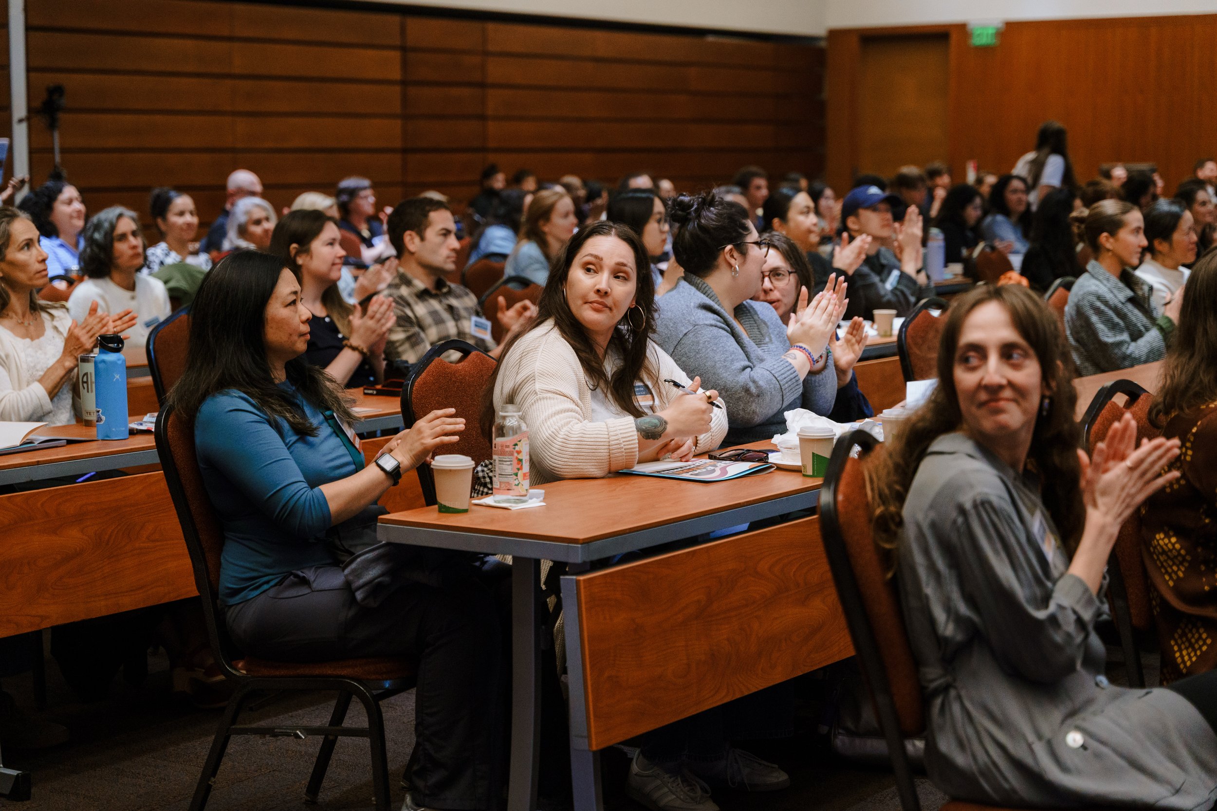 A large crowd of people attending a conference or seminar in a room with wooden paneling, sitting at long tables, some clapping and some taking notes.