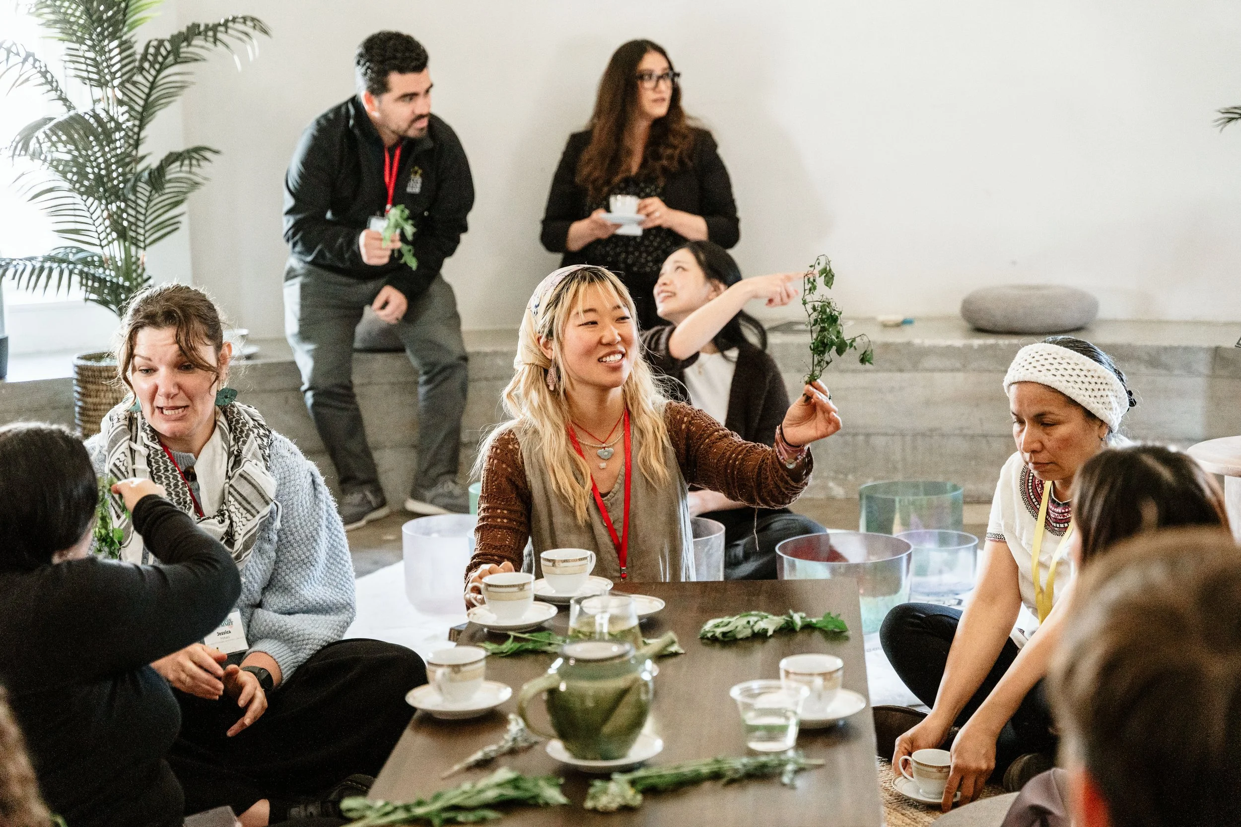Group of diverse women sitting on the floor and a woman standing, engaging in a workshop or discussion, with tea cups and plants on the table.