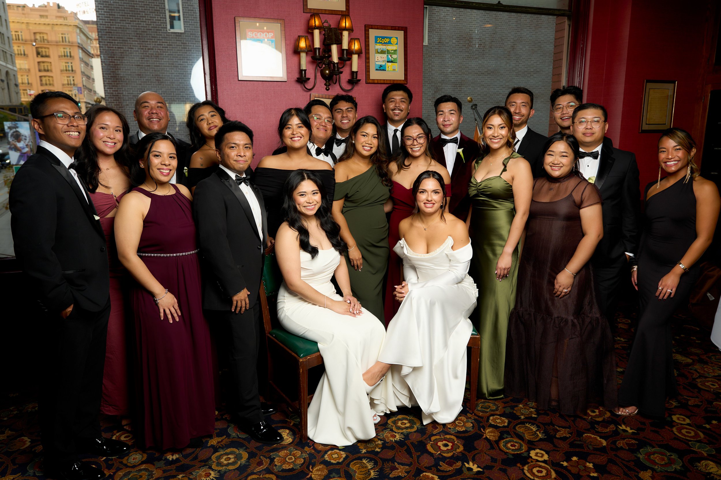 A group of diverse young adults dressed in formal attire at a wedding portrait booth, smiling and posing together indoors with a pink and brick wall background, framed pictures, and traditional wall lamps in San Francisco.
