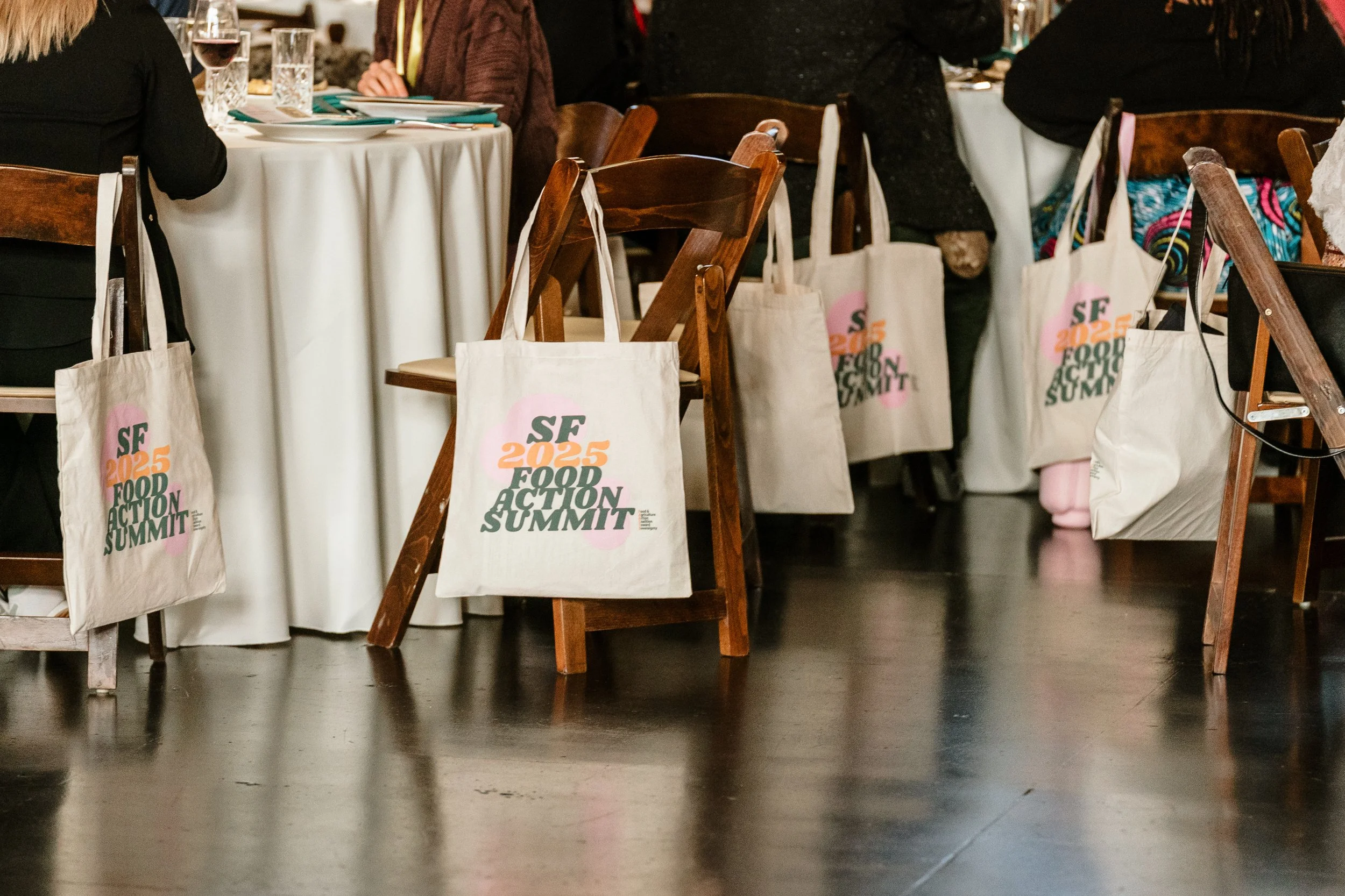 Several tote bags hanging on wooden chairs, with a white round dining table set with glasses and plates in the background at an event or gathering.