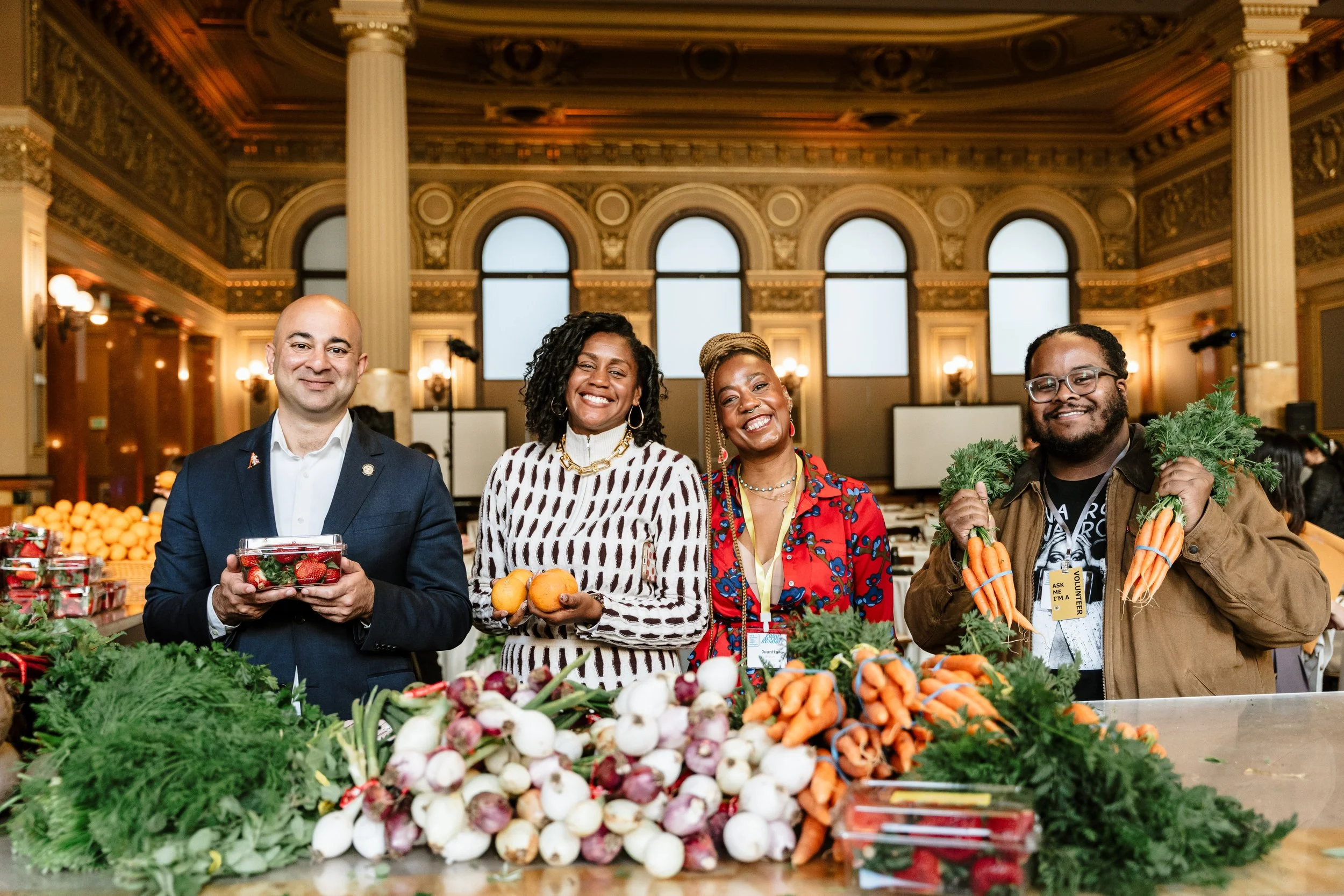 Four diverse people smiling and holding fresh vegetables at a farmers market, with tables of produce including carrots, radishes, and greens in front of them, inside a grand hall with tall windows and ornate gold detailing.