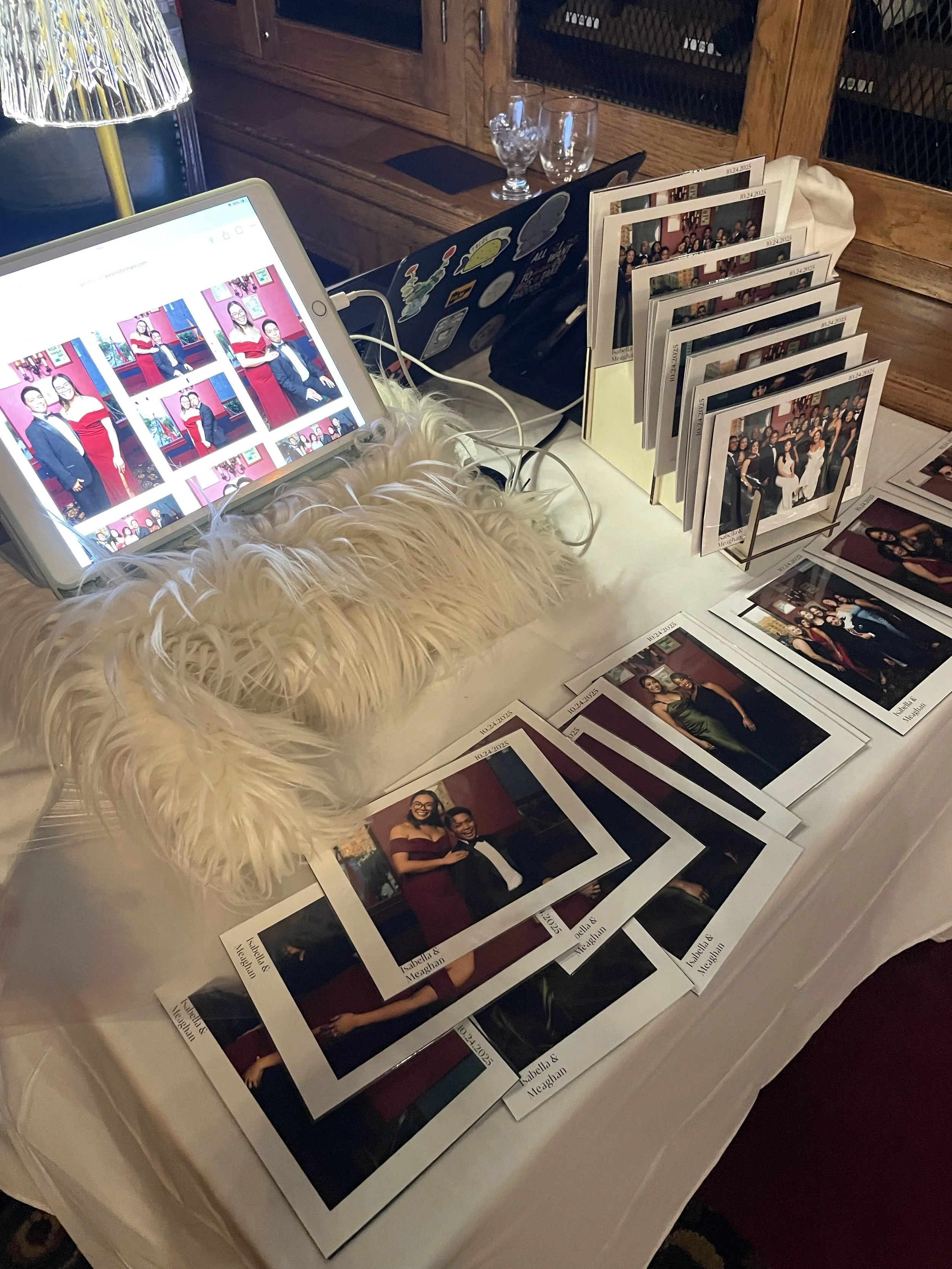 A look at a photography lounge station table with multiple printed photo cards, a white furry cloth, and a digital tablet displaying photos of people at a formal event. Behind the table, there are glassware and a wooden cabinet.