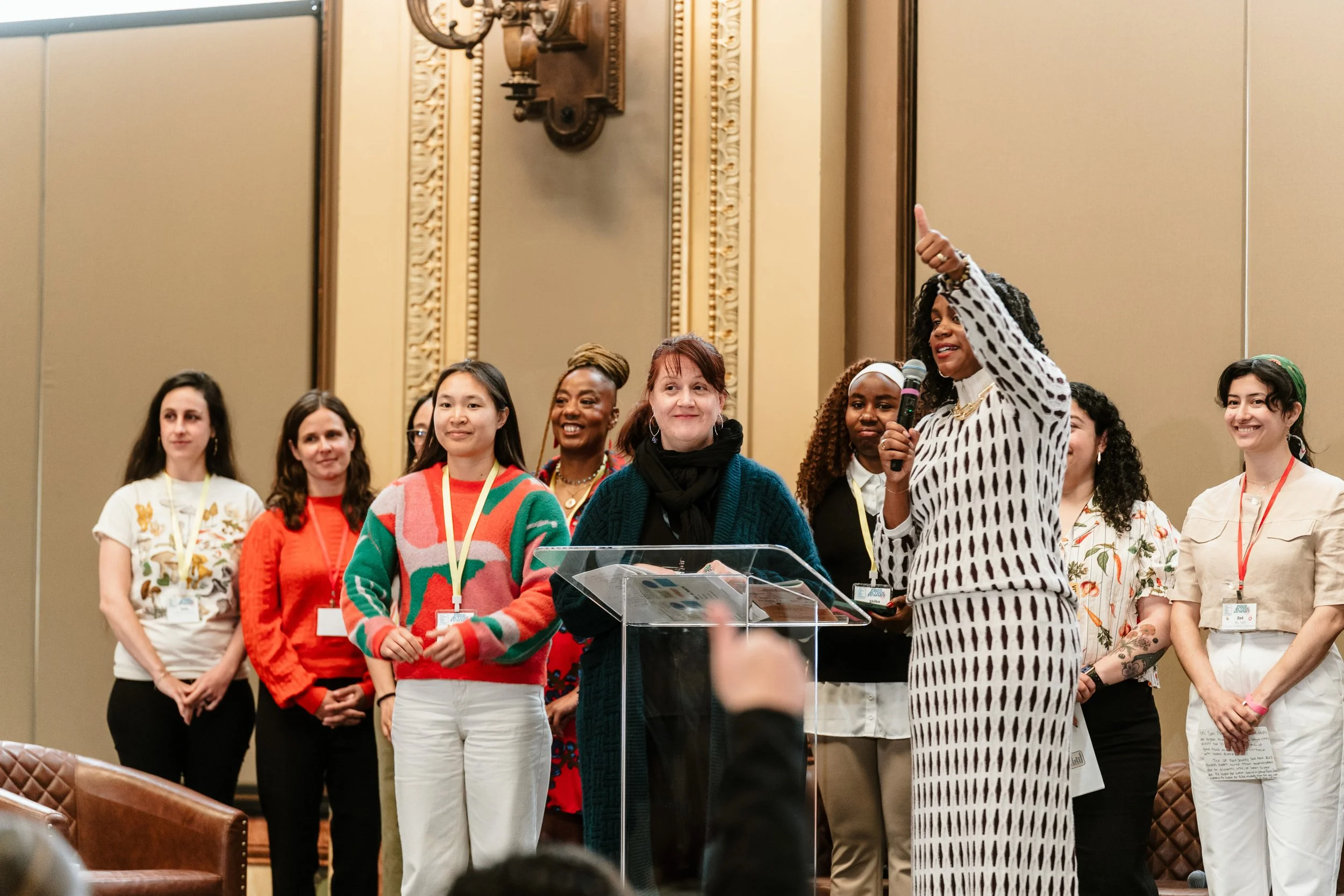 A diverse group of women standing on a stage during a conference or event, with one woman speaking into a microphone and gesturing. The women are dressed in colorful clothing and some are wearing name tags.