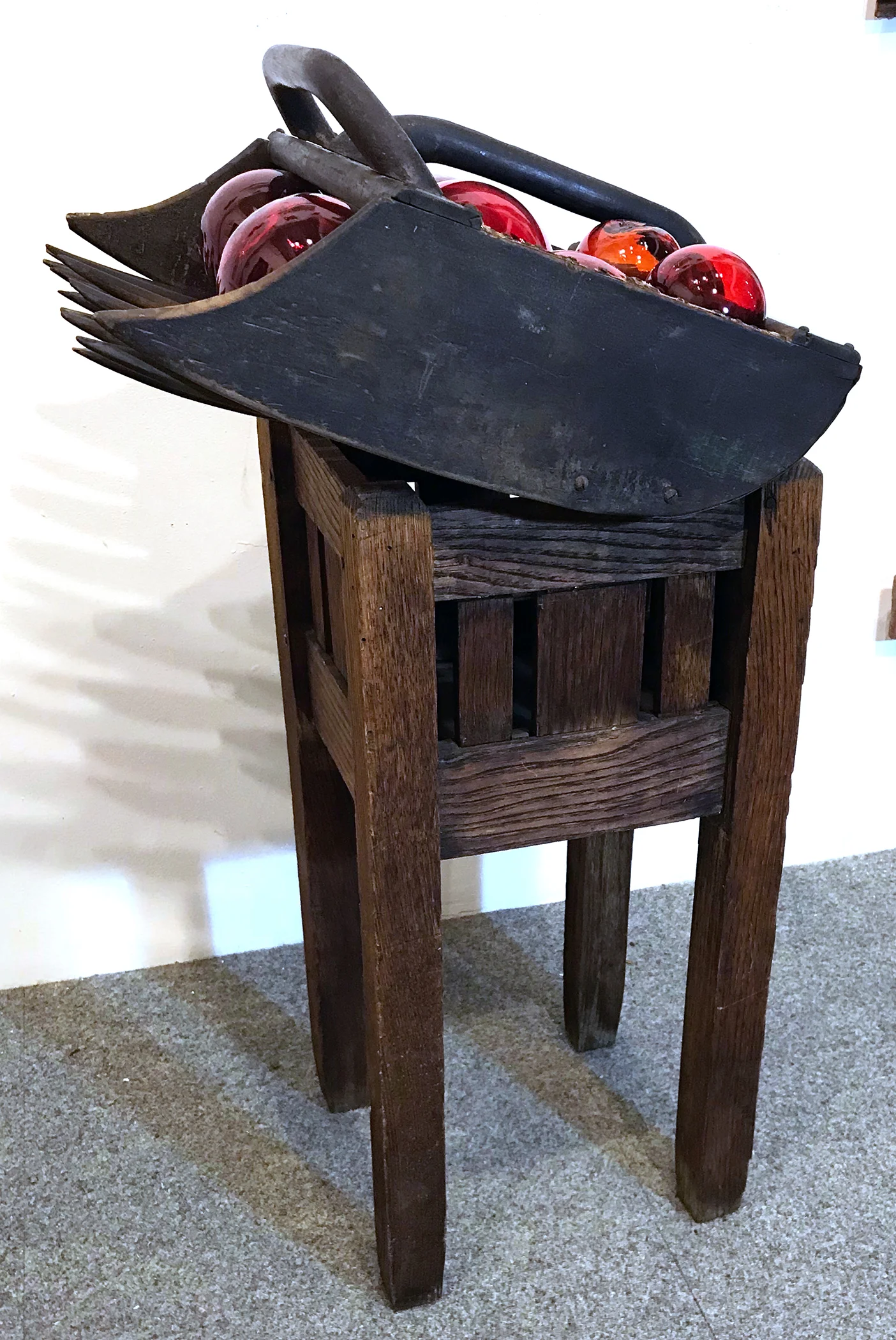 Cranberry. Assemblage of an antique cranberry rake filled with glass balls standing on an antique oak plant stand. Rake: 18x5x9"H. Stand: 12x12x25"H. 2018.