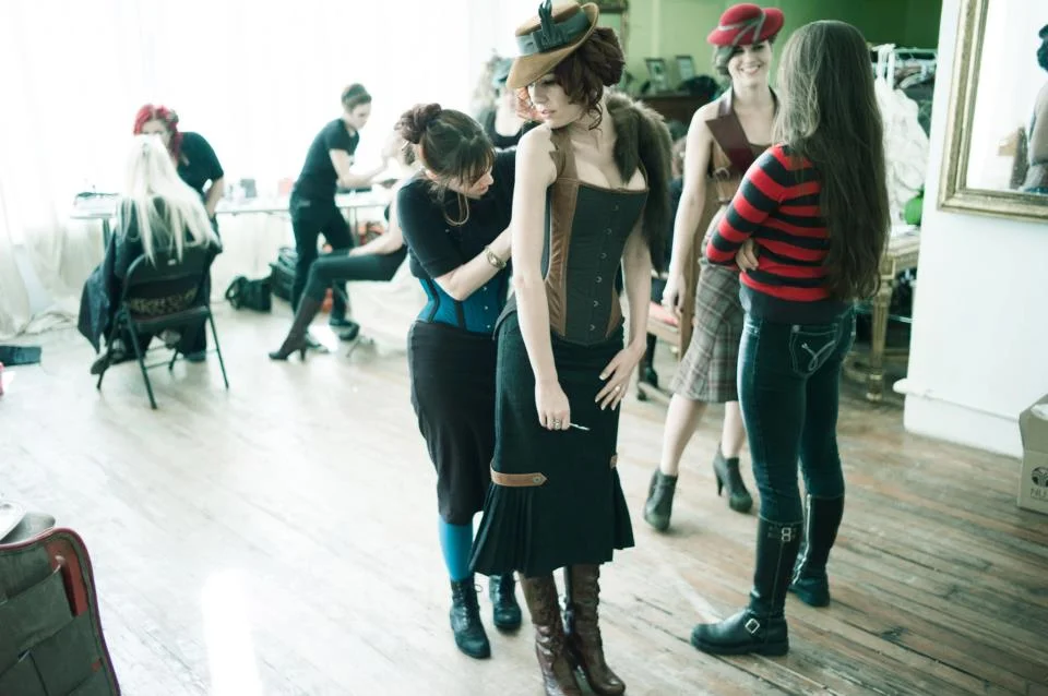 Behind the scenes at a Dark Garden photoshoot, Marianne (in a teal Waspie) laces in a model in a Personalized Design Dollymop Overbust . | Photo © Joel Aron