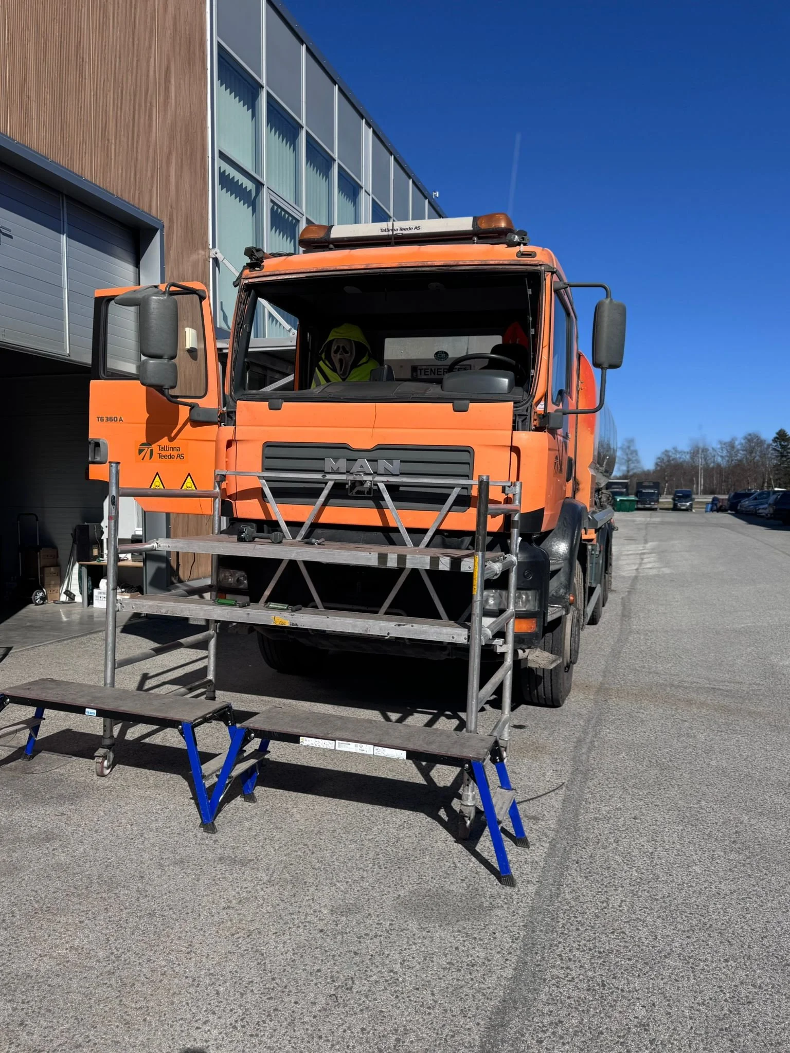 Orange construction truck parked outside a modern building under a clear blue sky, with scaffolding and blue steps in the foreground.