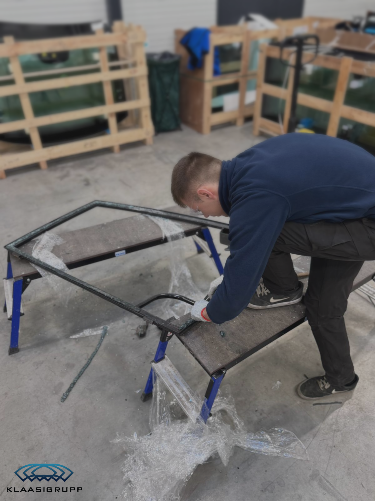 A person wearing glasses and gloves is working on a metal frame, possibly crafting or repairing, with plastic sheeting and tools on a work table. The setting appears to be a workshop or garage with wooden furniture and equipment in the background.