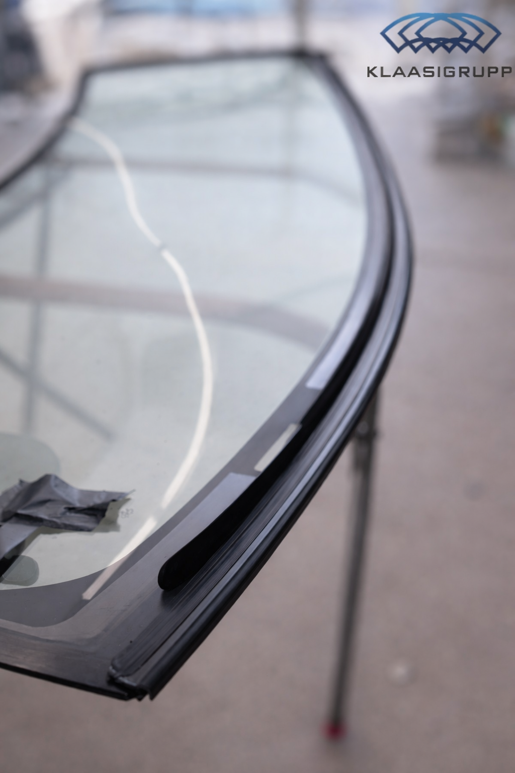 Close-up of a curved glass or plastic surface with black trim, possibly a windshield or display, with a blurred background and a logo that reads 'KLAASIGRUPP' in the top right corner.
