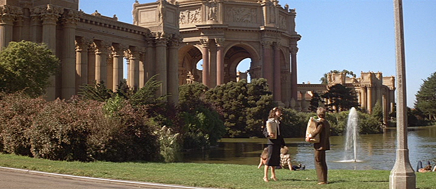 A man and a woman, both carrying bags of items, face each other in a lawn in front of a lake with a fountain and the Palace of Fine Arts.