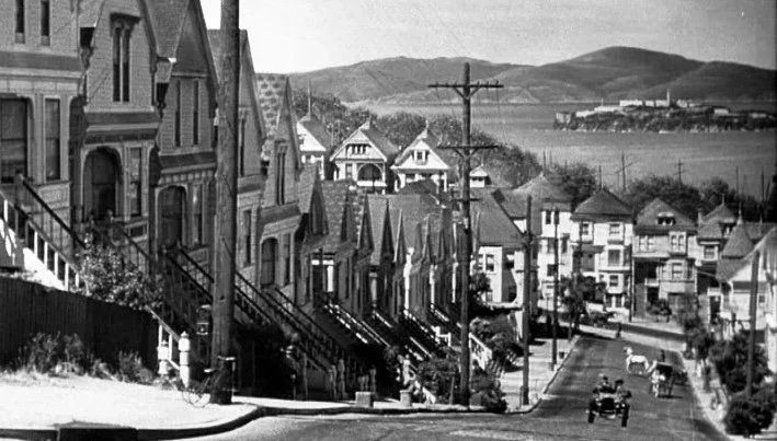 Black and white screenshot of a road on a hill with some Victorian houses on the left.