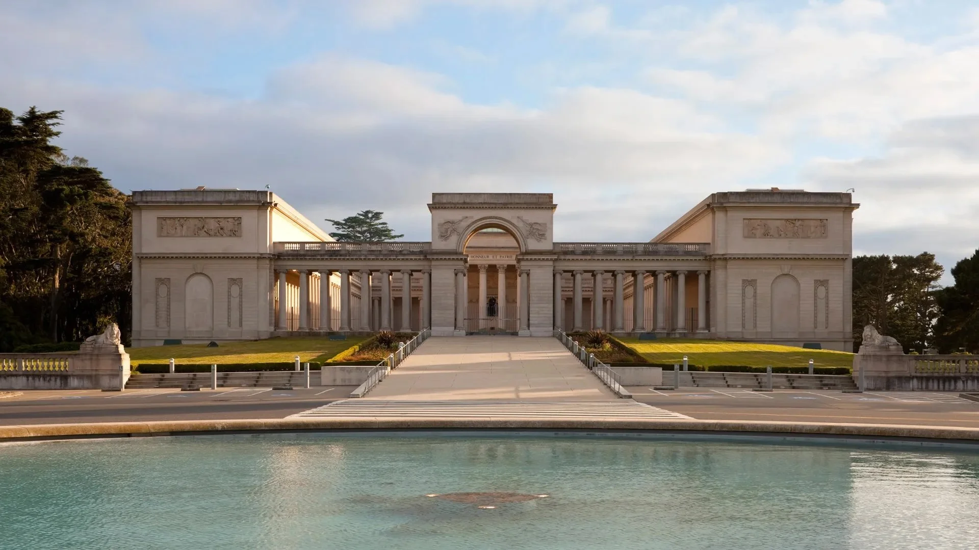 A long white building with Roman columns and an arch sits behind a lawn and a shallow pool of water.