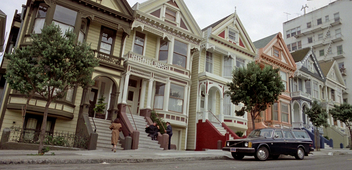 A woman walks up the steps to a yellow and brown Victorian house. There is a row of differently colored Victorian houses next to her.