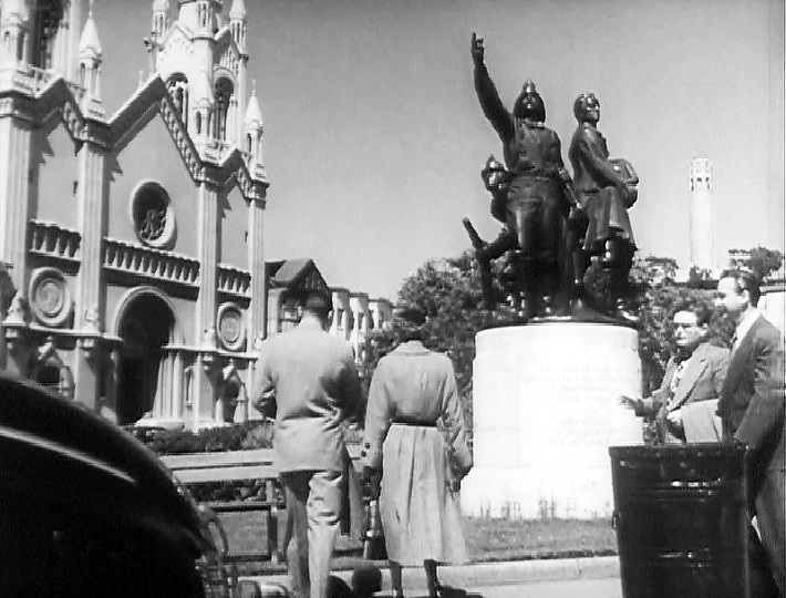 Black and white screenshot of a man and a woman walking towards a church and a statue with three soldiers on it.
