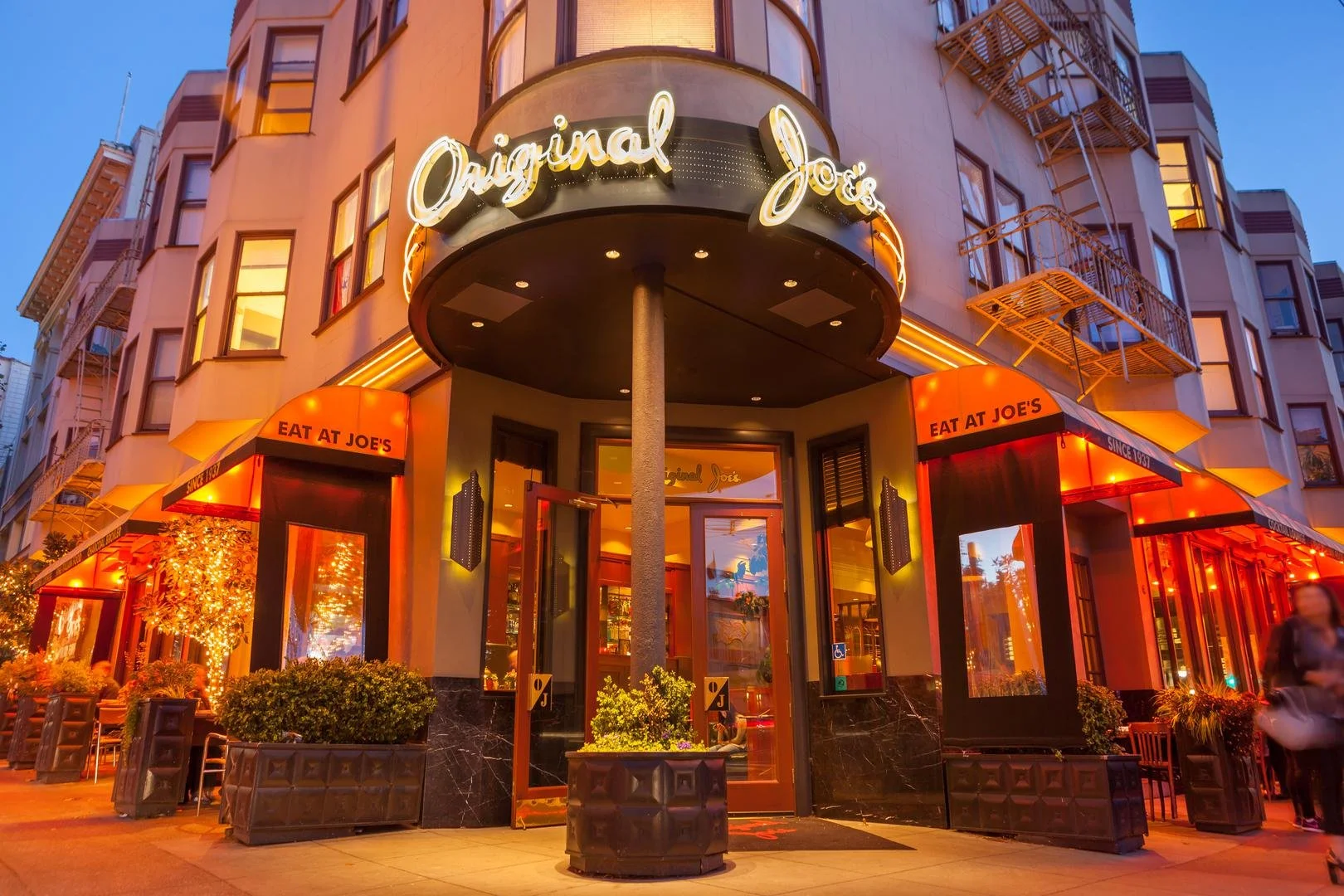 A restaurant entrance on a city corner with windows lit up red and overhangs reading "Eat at Joe's." The sign over the entrance says "Original Joe's" in neon white light.
