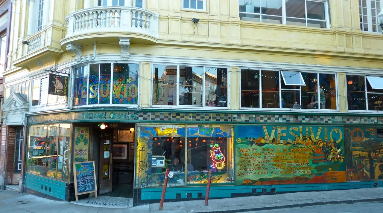 A green and blue tiled and painted wall spans a street corner. The sign above the door to the bar reads "Vesuvio" and is clearly painted on the inside of the window.