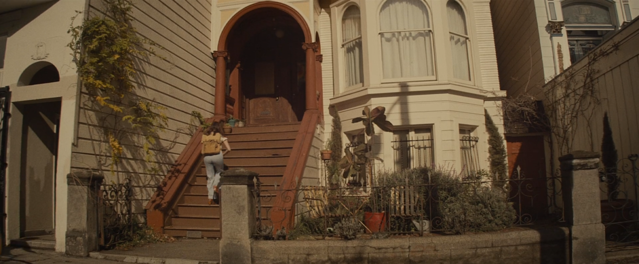 Screenshot of a teenage girl from the back wearing a beige backpack as she walks up some red stairs into a Victorian house.