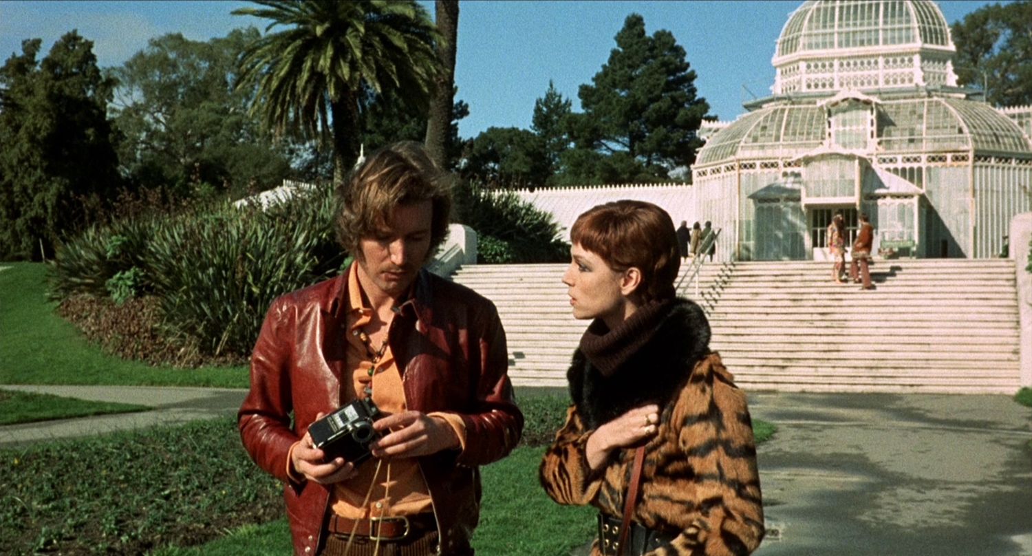 A man and a woman look at a camera while standing in the lawn in front of a white, wood-and-glass building.