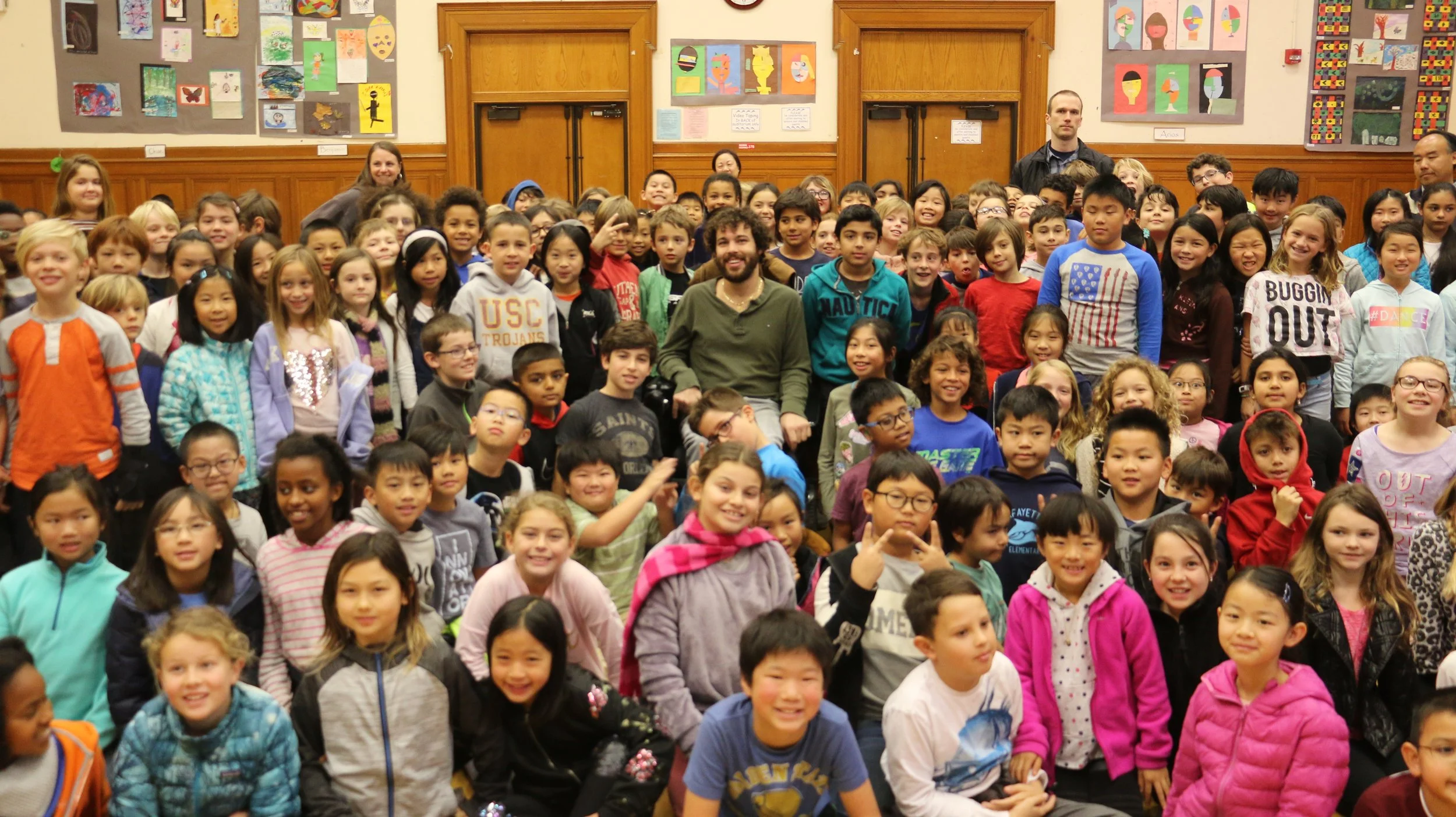 A hallway at a school is filled with young students around 7-10 years old. An adult man sits at their center and a few teachers are standing at the back. There is student art on the walls behind them.