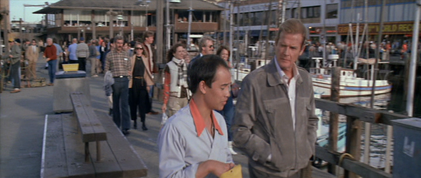 Two men walk along a crowded boardwalk with sailboats in the water next to them.